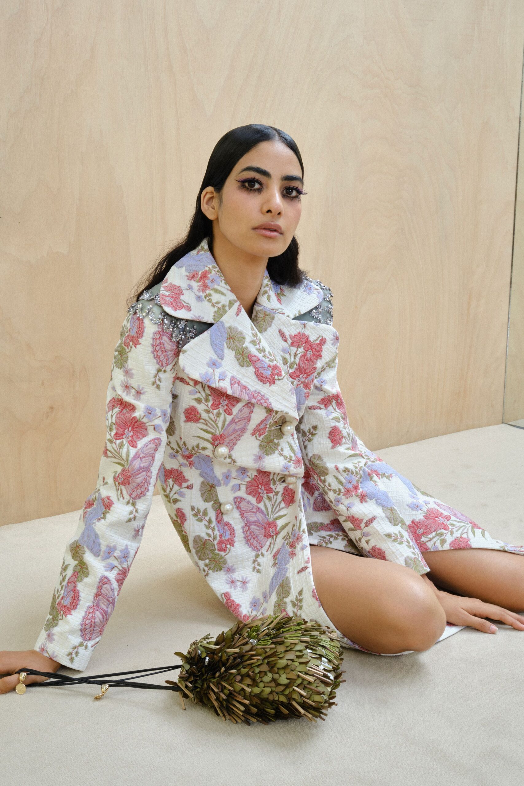 White matelasse coatdress printed with pink butterflies and olive vines, fastened in oversized pearls. Silver beaded epaulettes crusting the shoulder seams. Olive paillette pouch, bristling like seed pods on cords. Waiting on a plywood set as if the garden party got moved indoors.