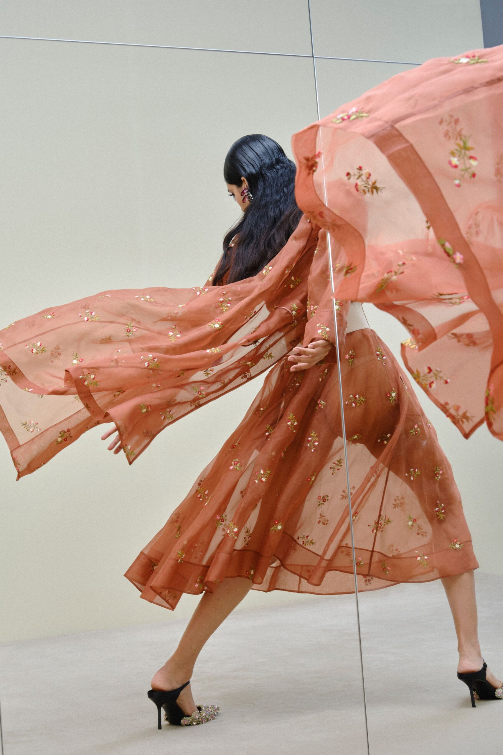 Burgundy drop earrings catching beside the slicked hairline. Cinnamon sheer organza dress scattered with tiny floral embroidery. Pale sash belt cinching the waist under the transparent layer. Black satin mules topped with clustered jeweled flowers. Crossing a hotel atrium when the revolving door sends everything airborne.