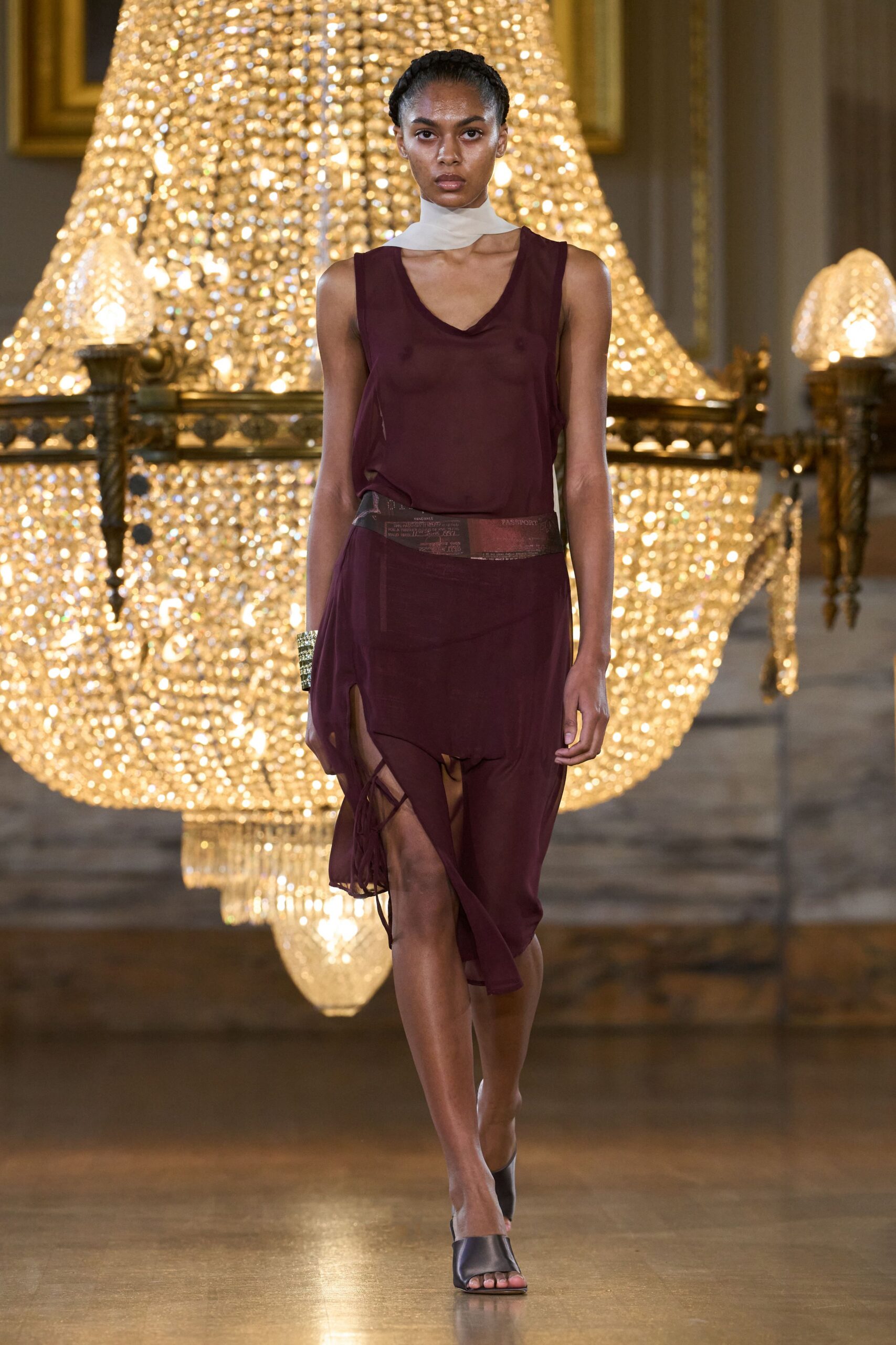 Chalk silk scarf wrapped close, one end crossing the throat. Burgundy sheer jersey tank dress, slit and laddered ties exposing one thigh. Transparent printed waistband sitting low across the hips. Black satin mules with a square peep toe. After midnight in a hotel ballroom, chandeliers still blazing.