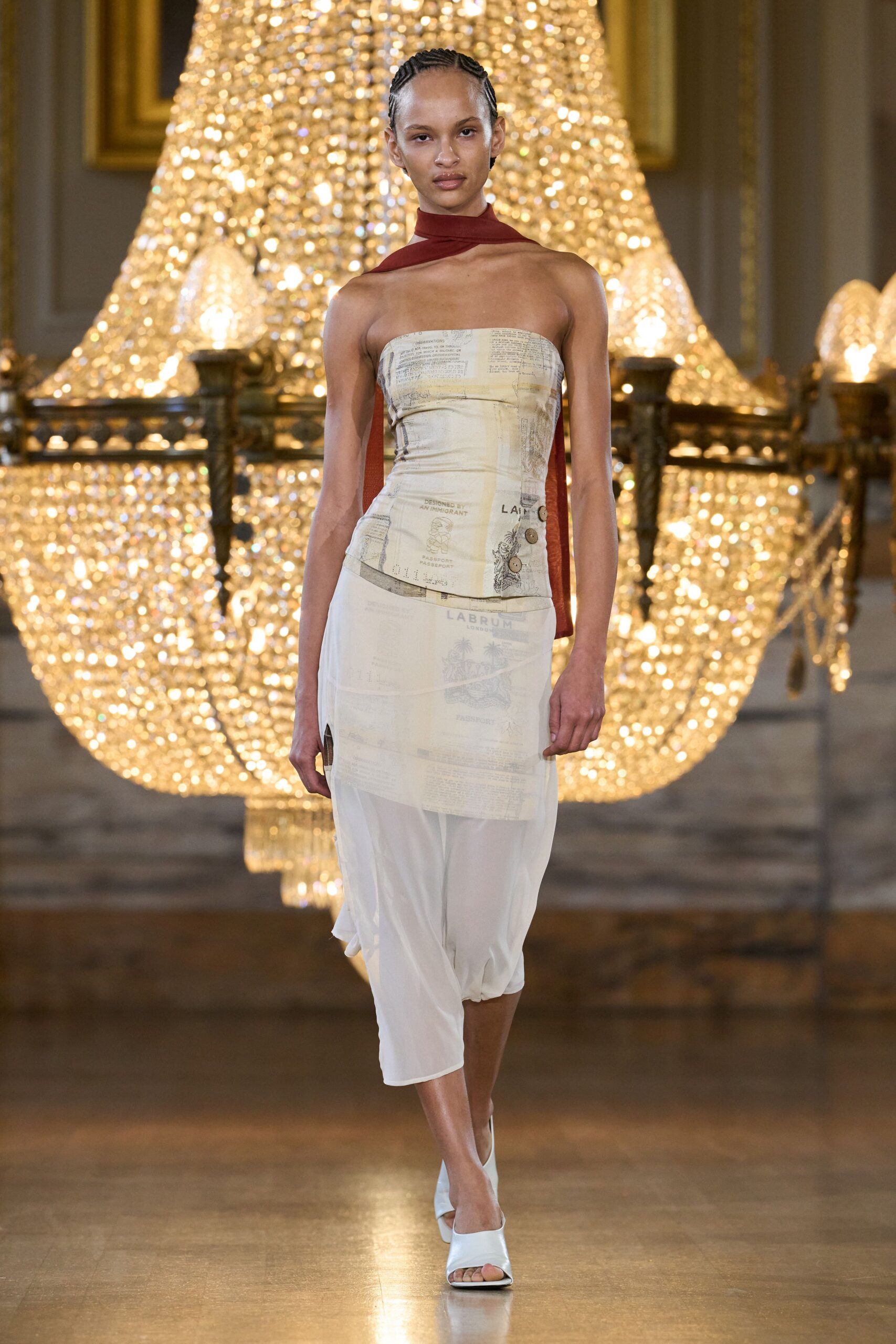 Rust jersey scarf crossing the throat, one tail dropped down the back. Strapless column in parchment mesh, overprinted with passport pages and side buttons. Sheer white cropped trousers, softly ballooned through the calf. White leather mules with a squared open toe. Crossing an arrivals hall still holding the paper customs form.