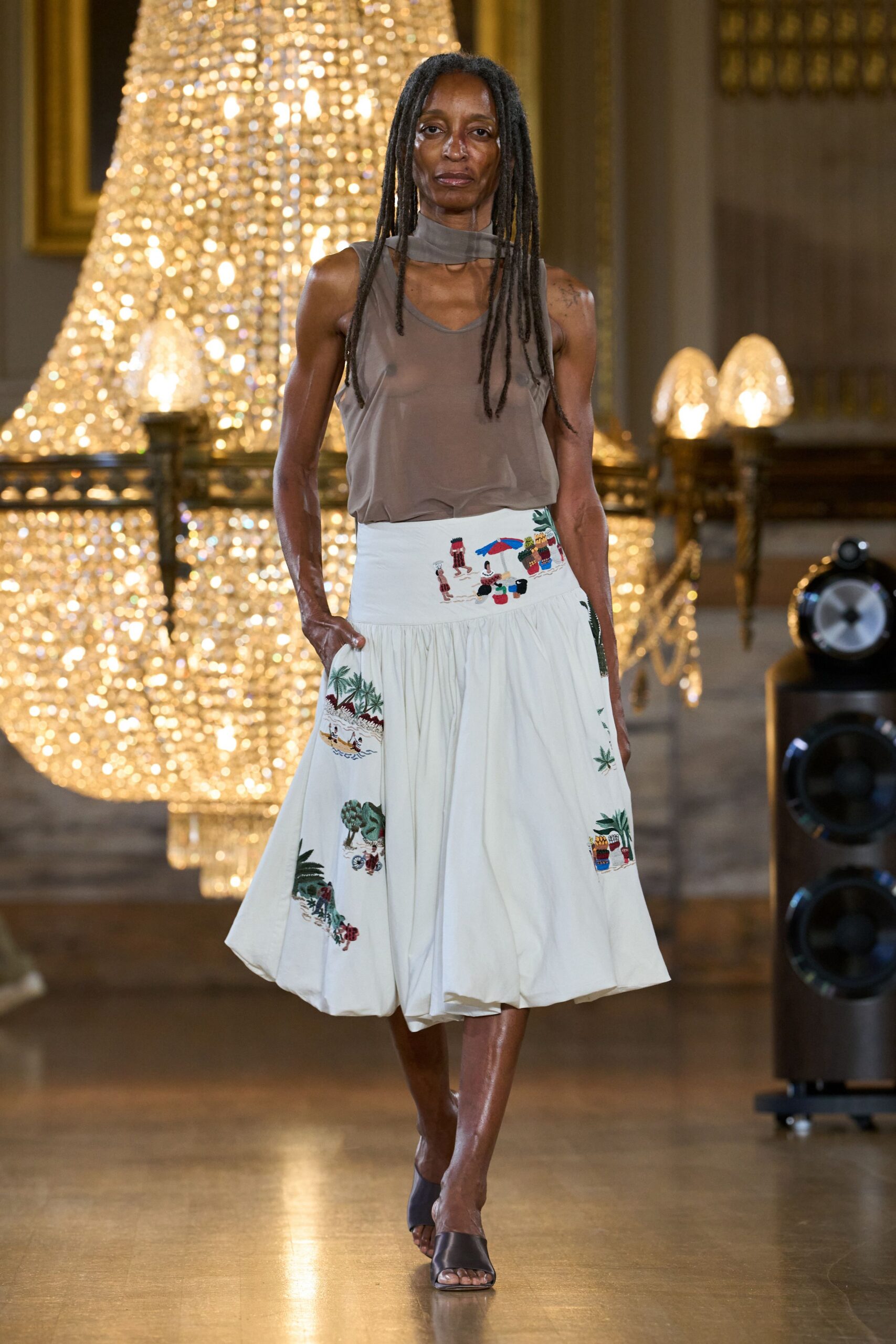 Taupe sheer jersey tank with a built-in scarf collar loop. White gathered midi skirt, hem puffed into a lantern shape, postcard embroidery at the waist and sides. Black leather mules with a broad vamp. Walking through a hotel ballroom after the wedding band has packed up.