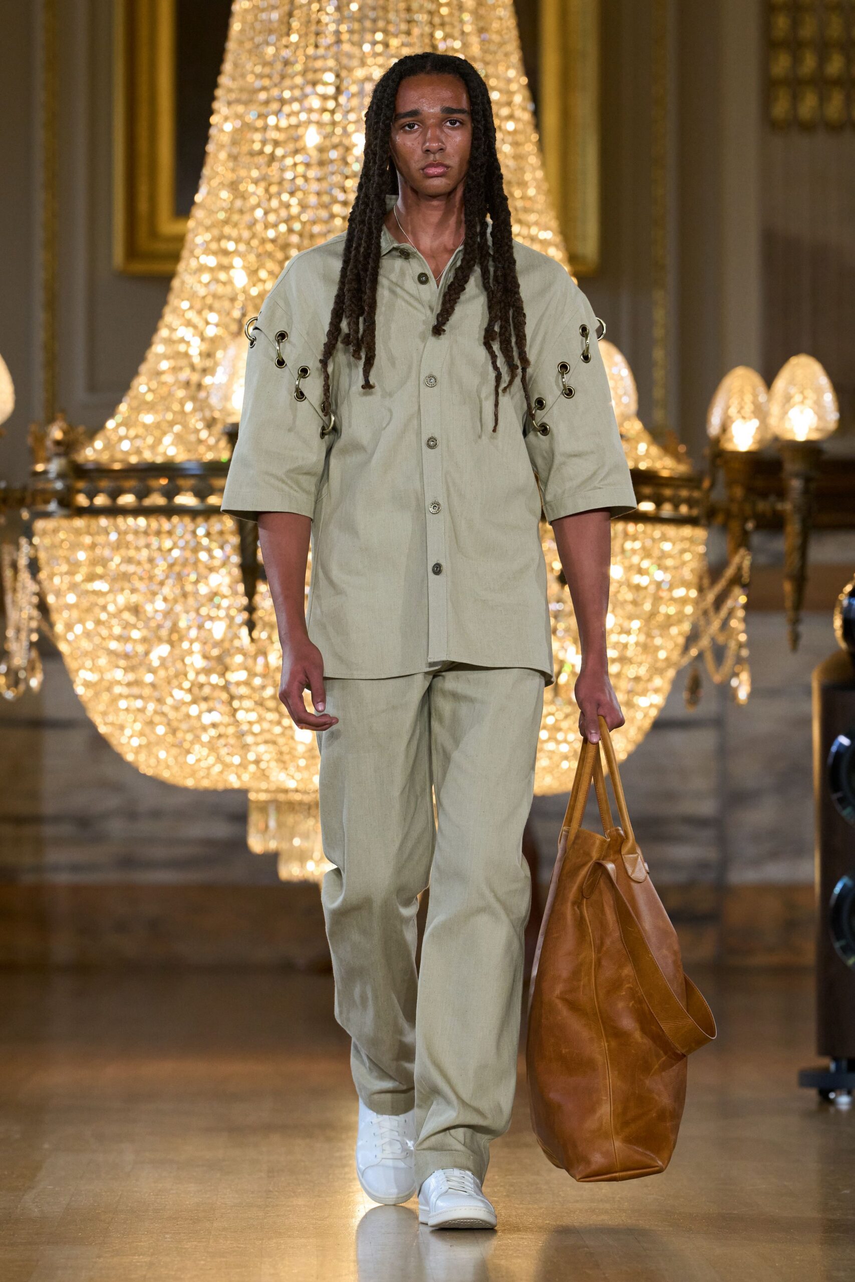 Pale sage short-sleeve shirt, sleeves split open with grommets and metal rings. Matching straight trousers in a dry linen-like weave. Oversized tan leather tote collapsing into soft creases. White leather low-tops. Walking into a country house after buying too much at the farm stand.