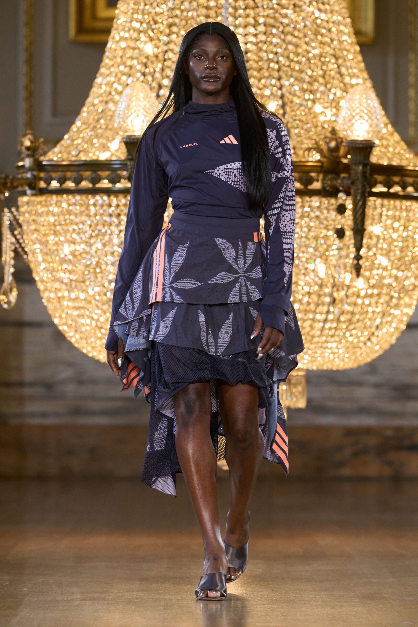 Navy stretch top with a ruched funnel neck and raglan seams. Asymmetric layered skirt in perforated mesh, grey leaf print and coral stripes. Gunmetal leather mules with a squared open toe. Walking fast through a museum atrium after the charity 5K.