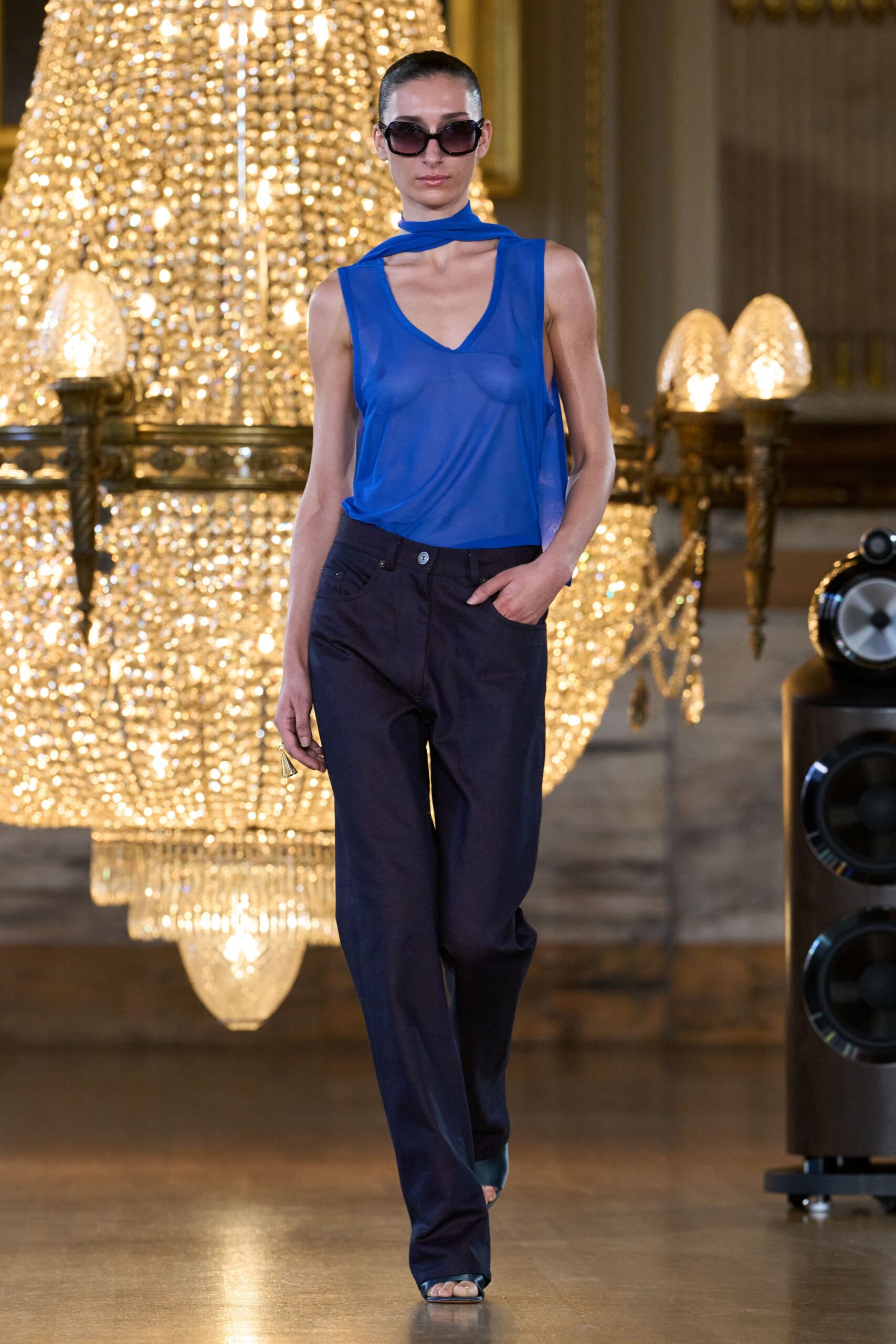 Oversized square frames with a smoke gradient lens. Cobalt sheer jersey tank with a scarf panel looped at the neck. Ink straight-leg trousers, silver shank button catching at the waistband. Blue satin slide sandals with a narrow open toe. Crossing a hotel ballroom after midnight, chandeliers still blazing.