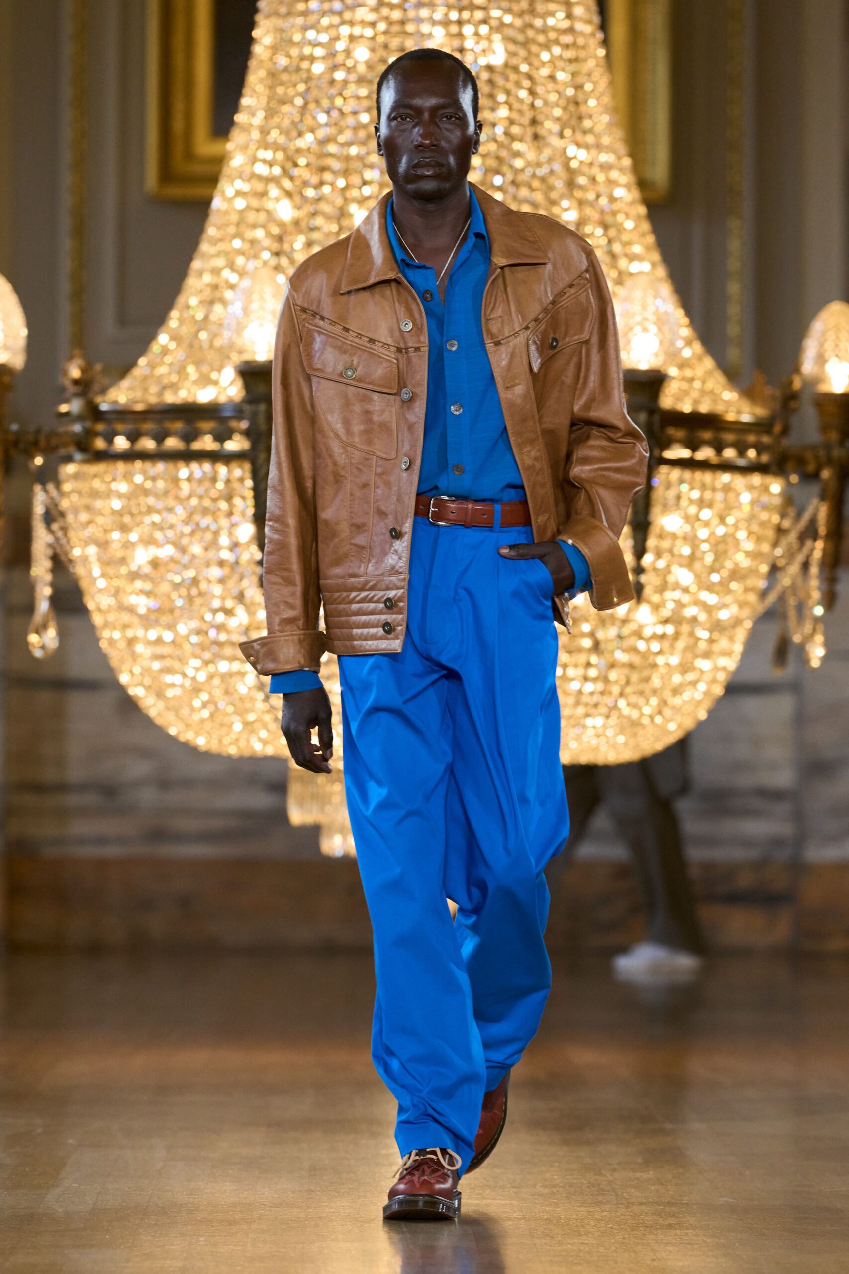 Tan leather trucker jacket with curved yoke seaming and ribbed side tabs. Electric-blue shirt, open collar under the jacket. Matching blue trousers falling loose from a brown leather belt. Oxblood lace-ups with pale piping at the throat. Walking through a chandelier-lit hall after changing out of black tie.