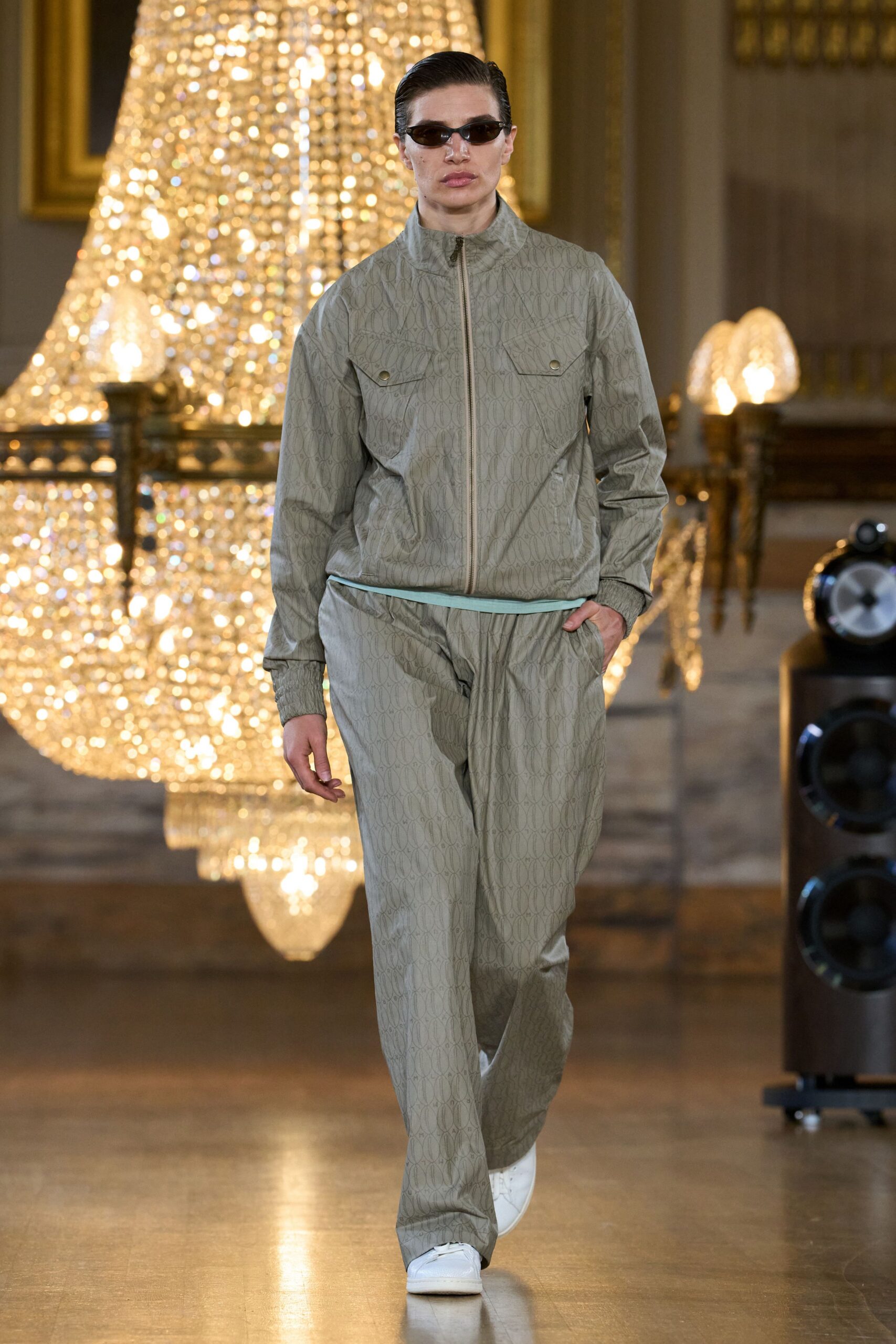 Narrow smoked wraparound sunglasses. Sage monogram track jacket, stand collar zipped high, snap-flap chest pockets. Mint crewneck peeking below the hem. Matching loose track pants in the same slick monogram weave. White leather sneakers, blunt and clean. Crossing a hotel ballroom before breakfast, chandeliers still burning.
