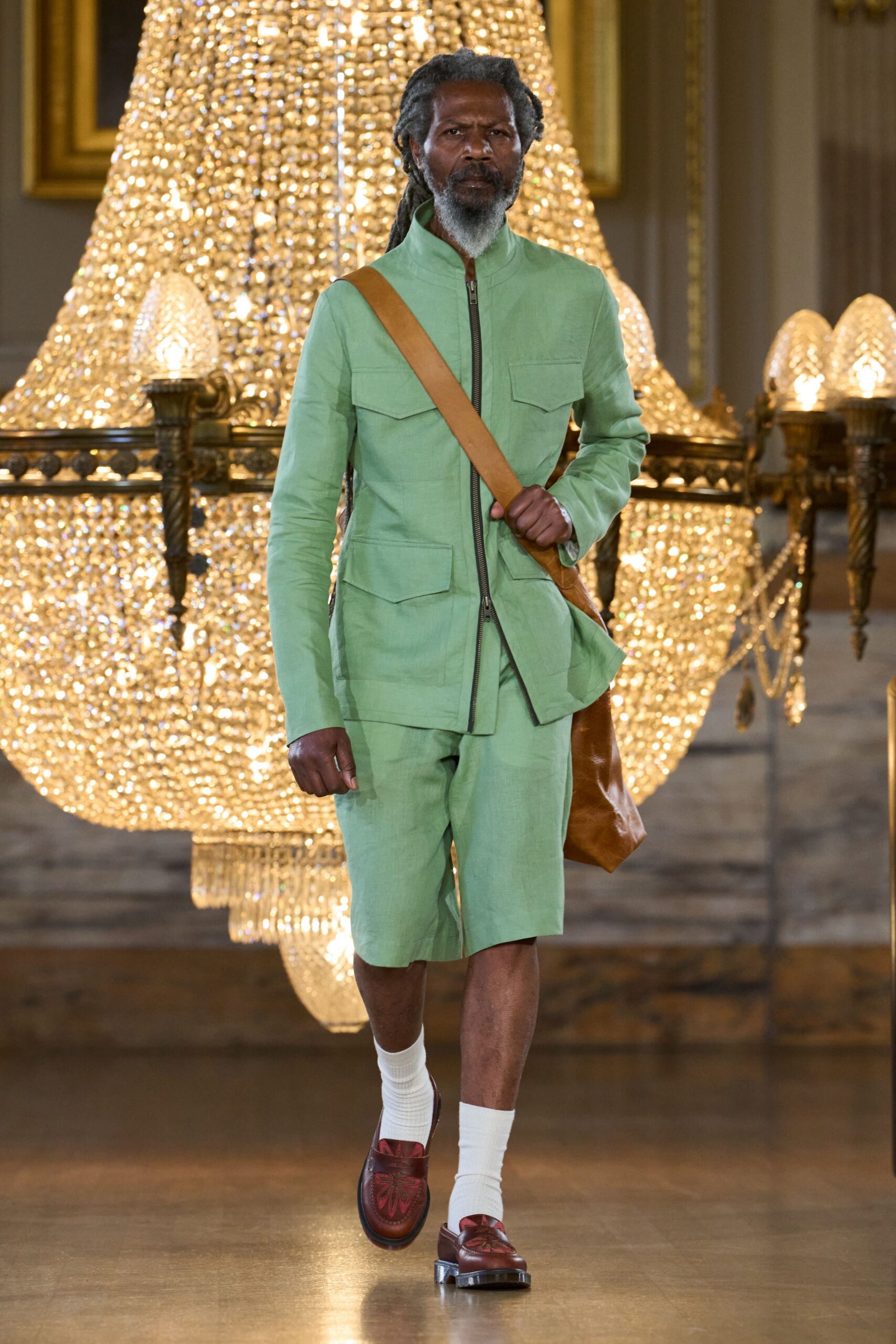 Mint linen field jacket, stand collar and four flap pockets. Matching knee-length linen shorts, softly creased through the thigh. Cognac leather satchel on a wide shoulder strap. White ribbed crew socks. Oxblood leather loafers with a dark floral vamp. Crossing a hotel ballroom early, before the wedding guests come down.