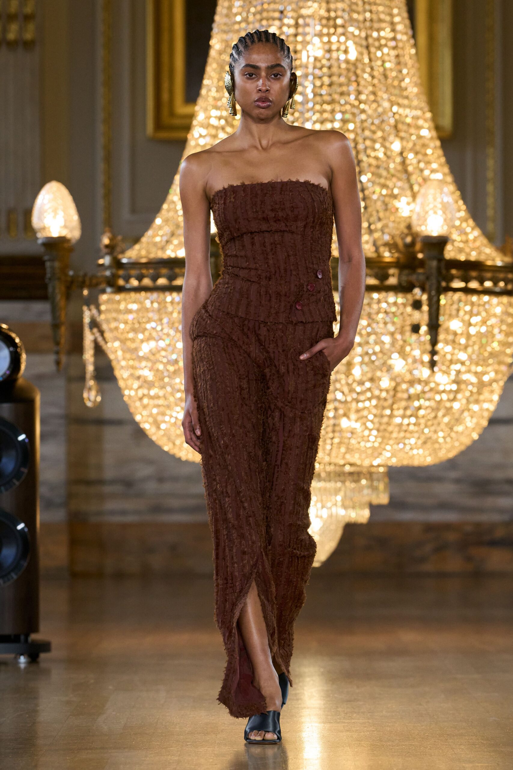 Hammered gold disc earrings with narrow drop pendants. Strapless espresso corset in frayed eyelash jacquard, three buttons off-center. Matching column skirt, twisted wrap front opening to the shin. Black leather peep-toe mules. Crossing a hotel ballroom after the speeches, chandeliers still blazing.