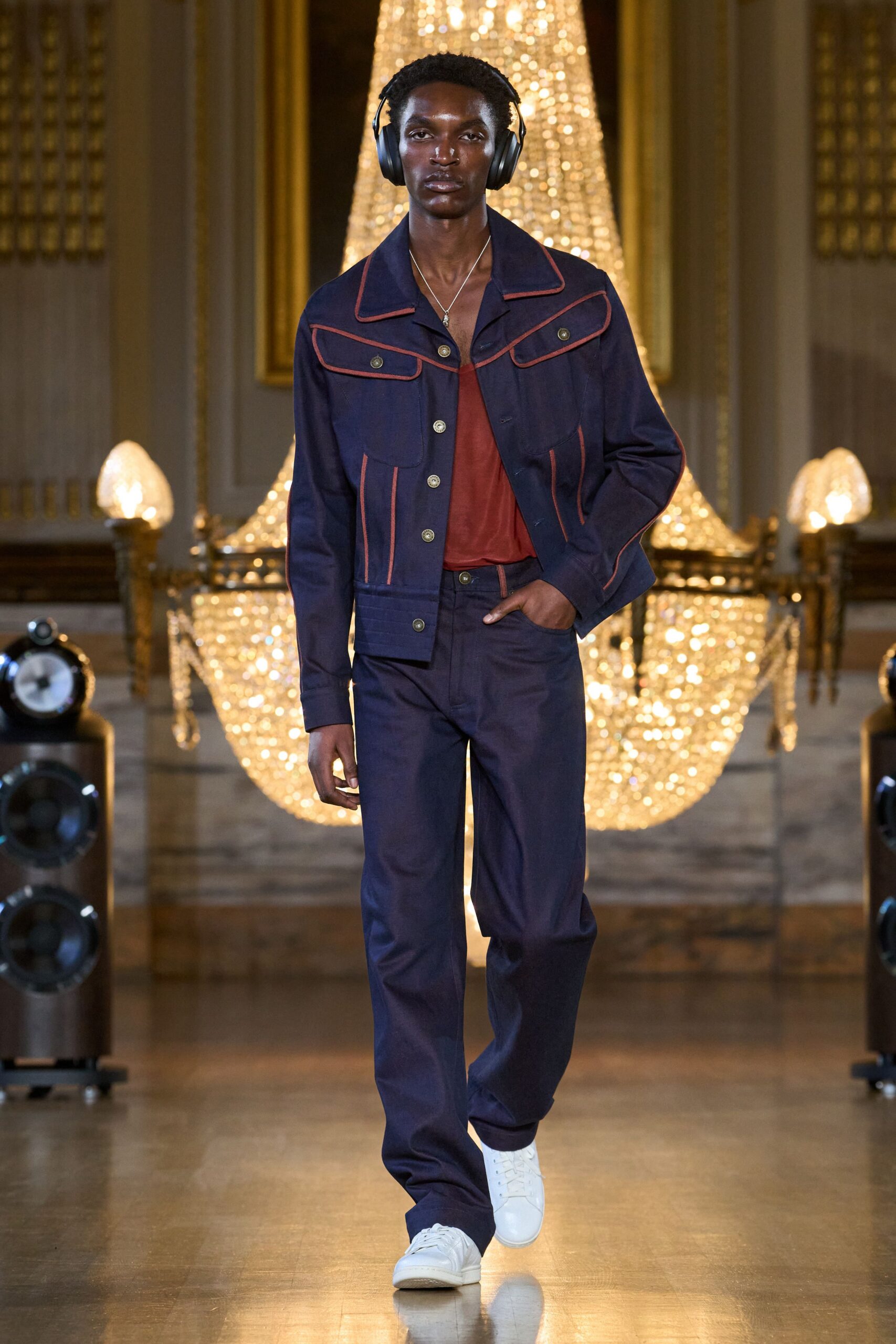 Black over-ear headphones worn as part of the look. Indigo workwear jacket traced with rust piping and oversized flap yoke. Brick satin camisole slumping open at the chest. Straight indigo trousers with matching rust side piping. White leather sneakers. Crossing a gilded ballroom before anyone notices the music changed.