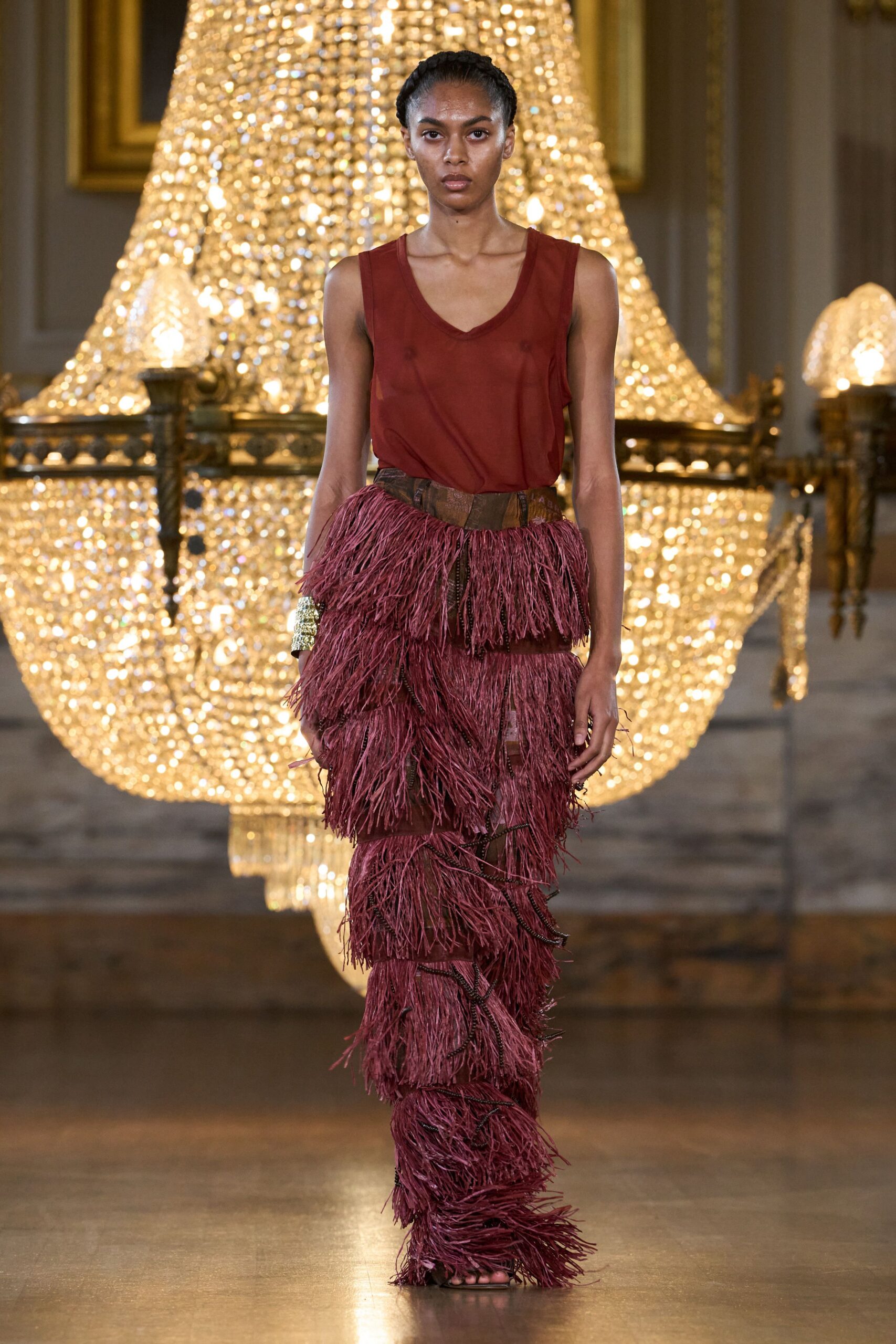 Sheer rust jersey tank with a deep scooped V neckline. Wine trousers buried under tiered raffia fringe, zip tape snaking between panels. Wide gold cuff, hammered and bright at one wrist. Dark barely there sandals peeking beneath the fringe. Crossing a chandeliered ballroom after midnight with tinsel from somewhere else.