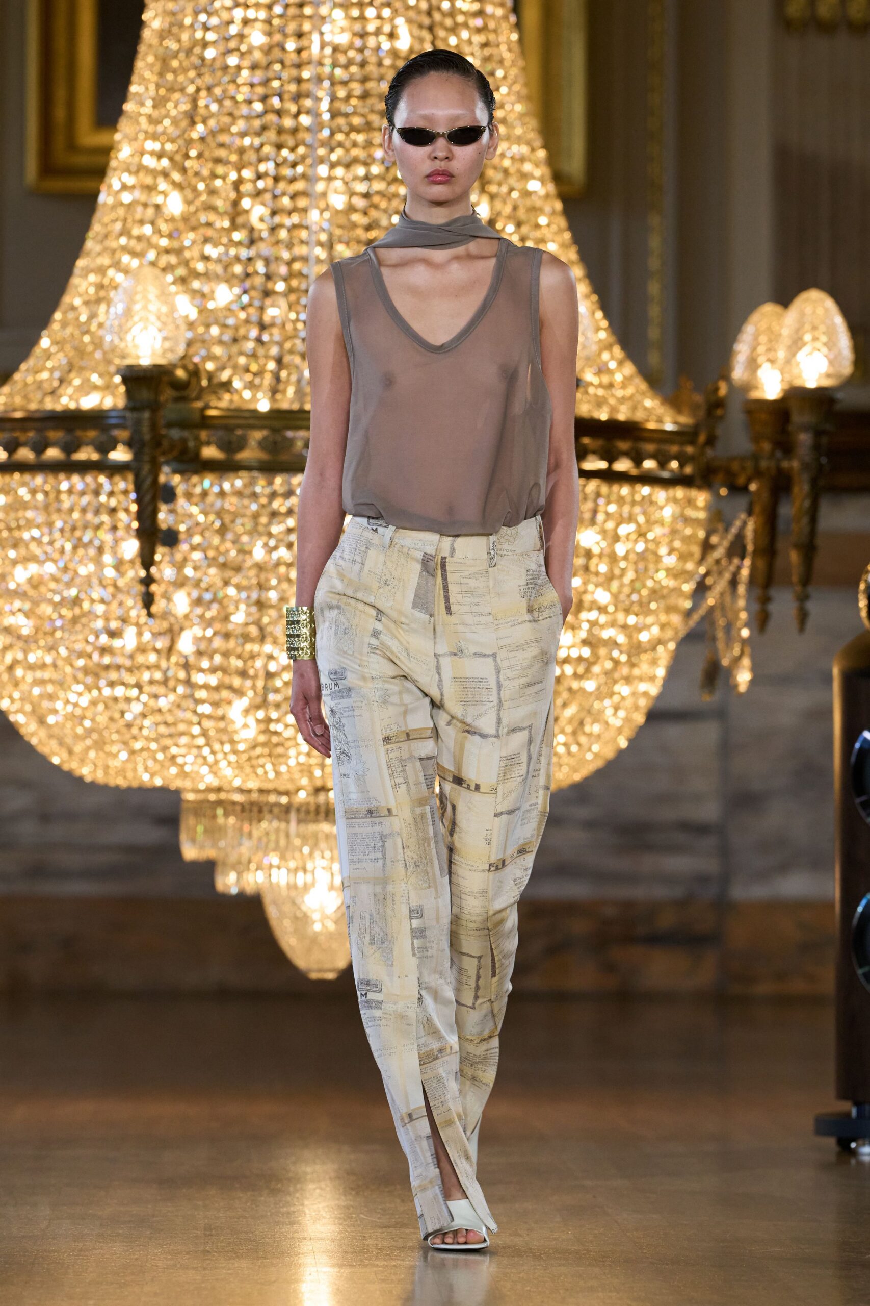 Narrow black oval sunglasses with thin pale rims. Taupe sheer jersey tank with a built-in neck wrap. Cream satin trousers printed like faded receipts, split over the toes. Wide gold cuff in stacked crystal rows. White square-toe sandals. Walking through a museum ballroom before the cleaning crew arrives.