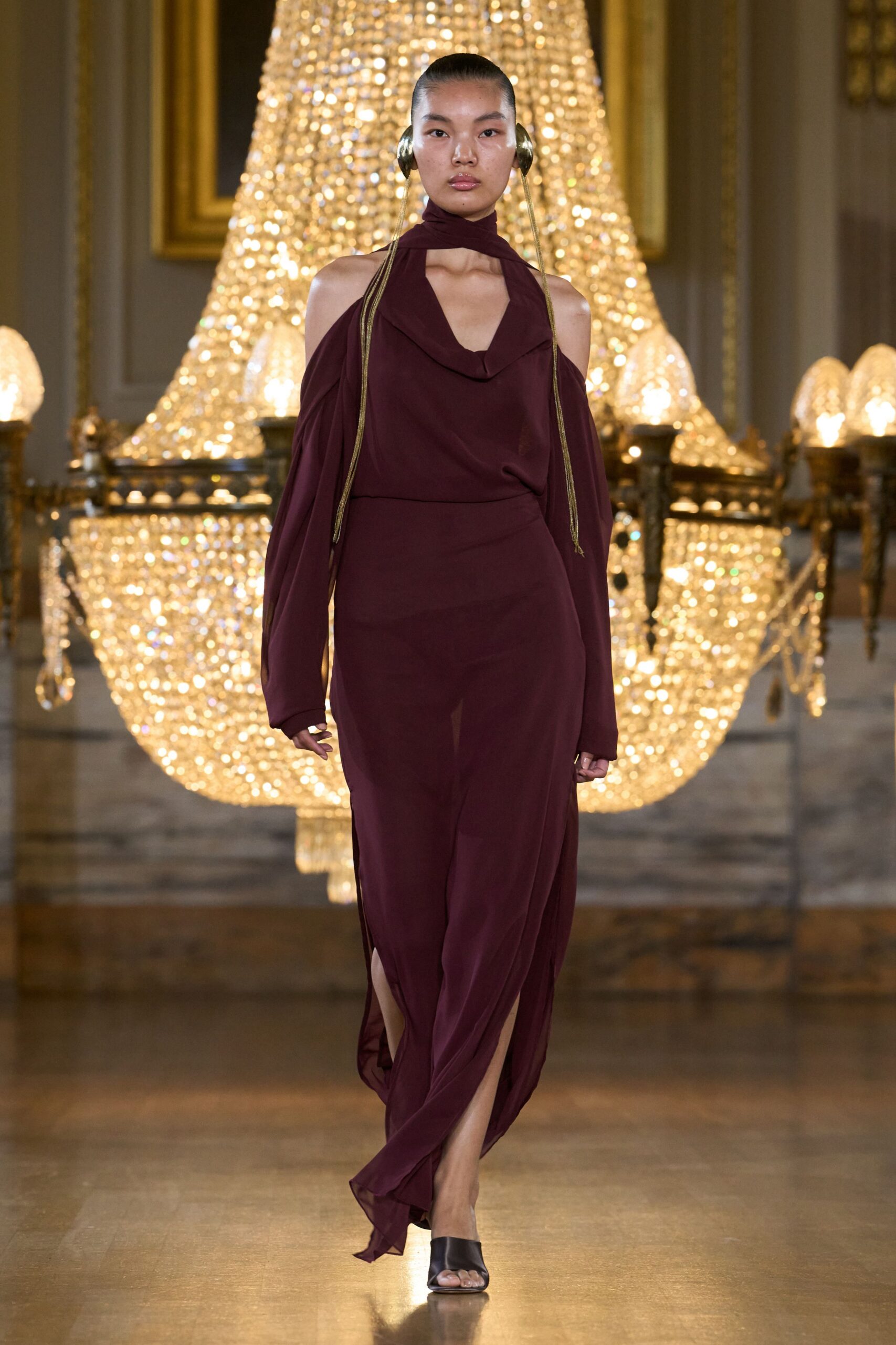Burgundy jersey dress with a scarf neck, cowl front and bare shoulders. Detached bishop sleeves hanging long over the hands. Brass orb earpieces with rope cords trailing to the waist. Black leather mule sandals, square at the toe. Crossing a ballroom floor before anyone notices the music stopped.