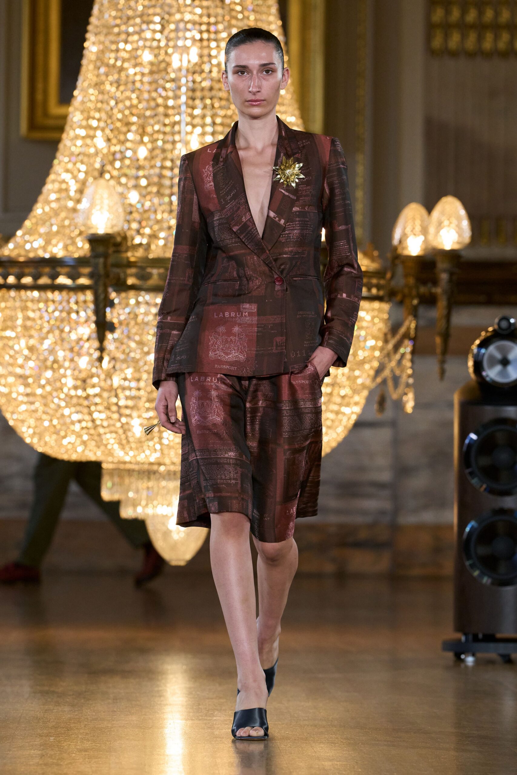 Deep-plunge blazer in burgundy satin, overprinted with passport pages and seals. Gold starburst brooch pinned high on the lapel. Bermuda shorts in the same glossy passport-print satin. Black leather mules with a squared open toe. Crossing a marble hotel lobby after a long-haul flight upgrade.