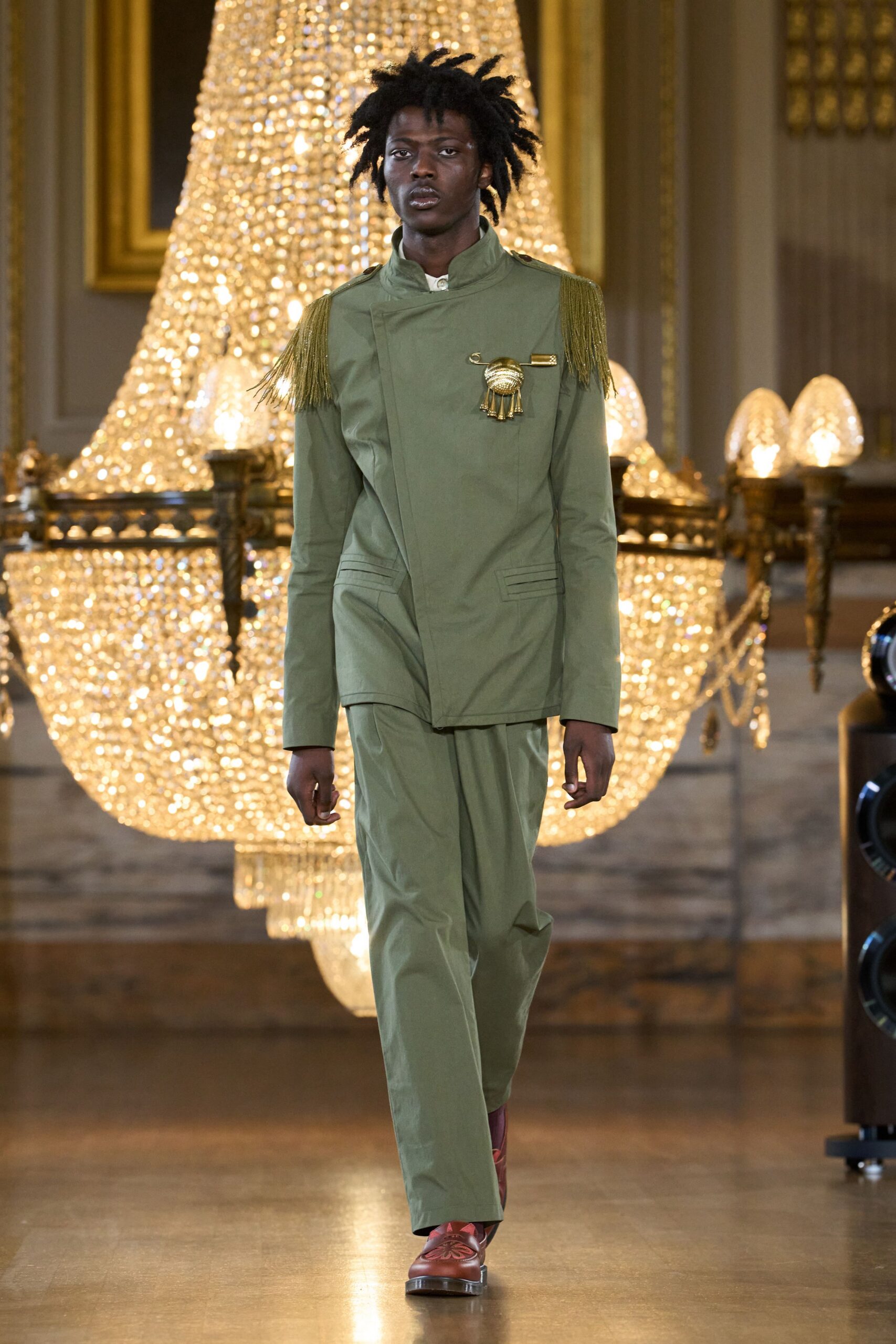 Sage drill jacket with an off-center closure, bullion fringe on both shoulders. Oversized gold safety-pin brooch with a tasselled medallion. Matching straight trousers in the same crisp sage cloth. Oxblood leather loafers with a cutout flower on the vamp. Crossing a palace ballroom after midnight, still in borrowed uniform.