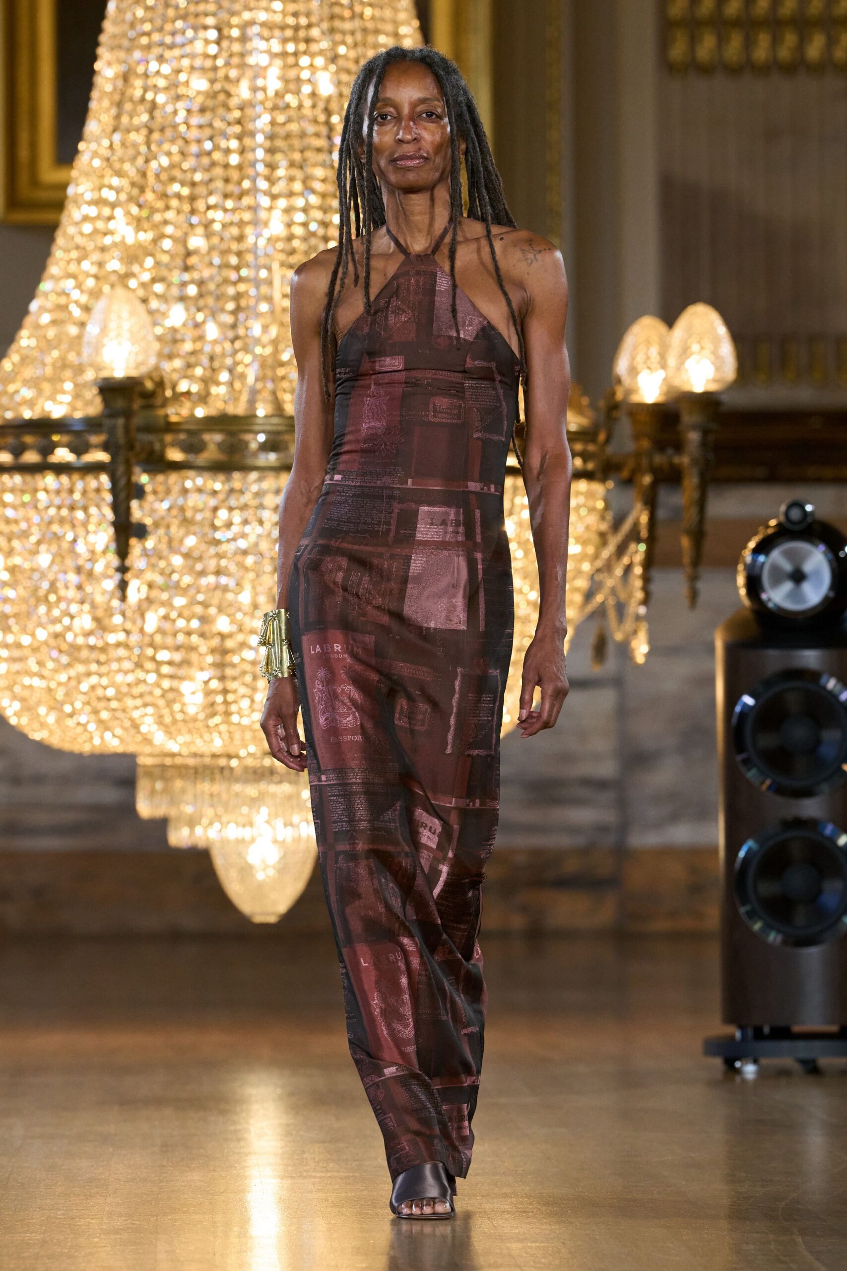 Burgundy mesh halter jumpsuit, collaged with passport pages and document blocks. Wide gold cuff with pleated fringe catching at the wrist. Black satin mule sandals, squared open toe. Under a chandelier after midnight, still carrying your papers.