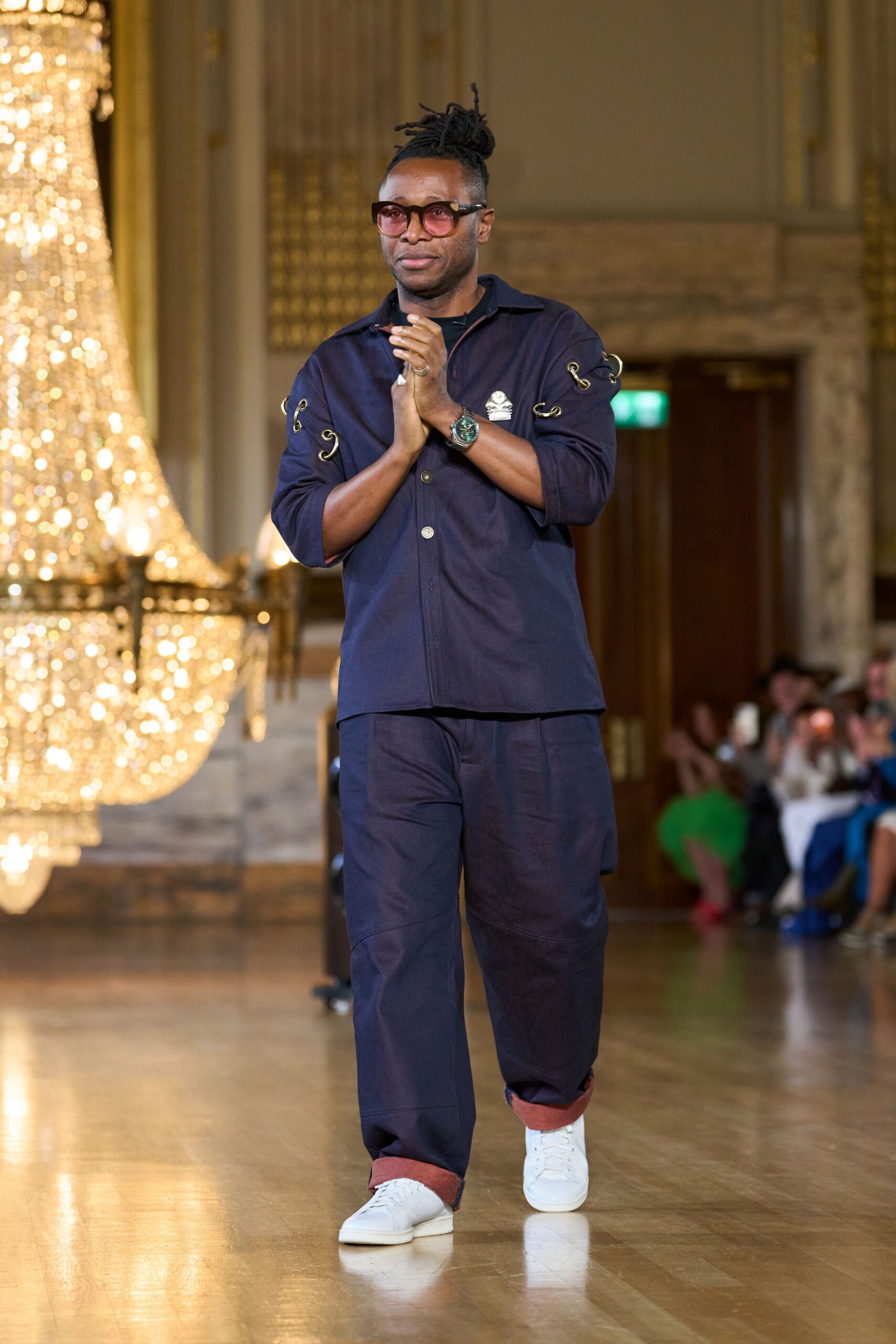 Rose-tinted square frames. Deep navy camp-collar shirt, sleeves laced with brass grommet rings. Matching wide trousers with curved knee seams and rust-turnback cuffs. White leather low-tops. Applause under chandeliers after a community hall fundraiser runs late.