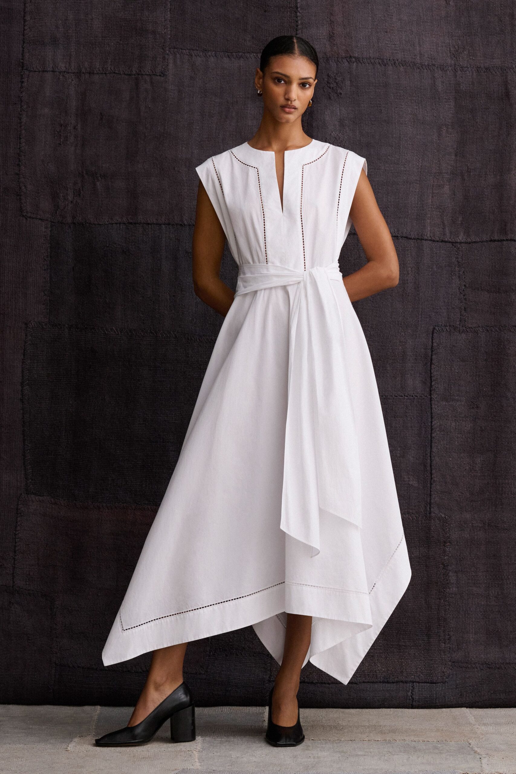 White cotton poplin dress with ladder-stitch seams and a long wrapped sash. Black leather pumps with squared toes and thick column heels. Crossing a sunstruck courtyard before the civil ceremony starts.