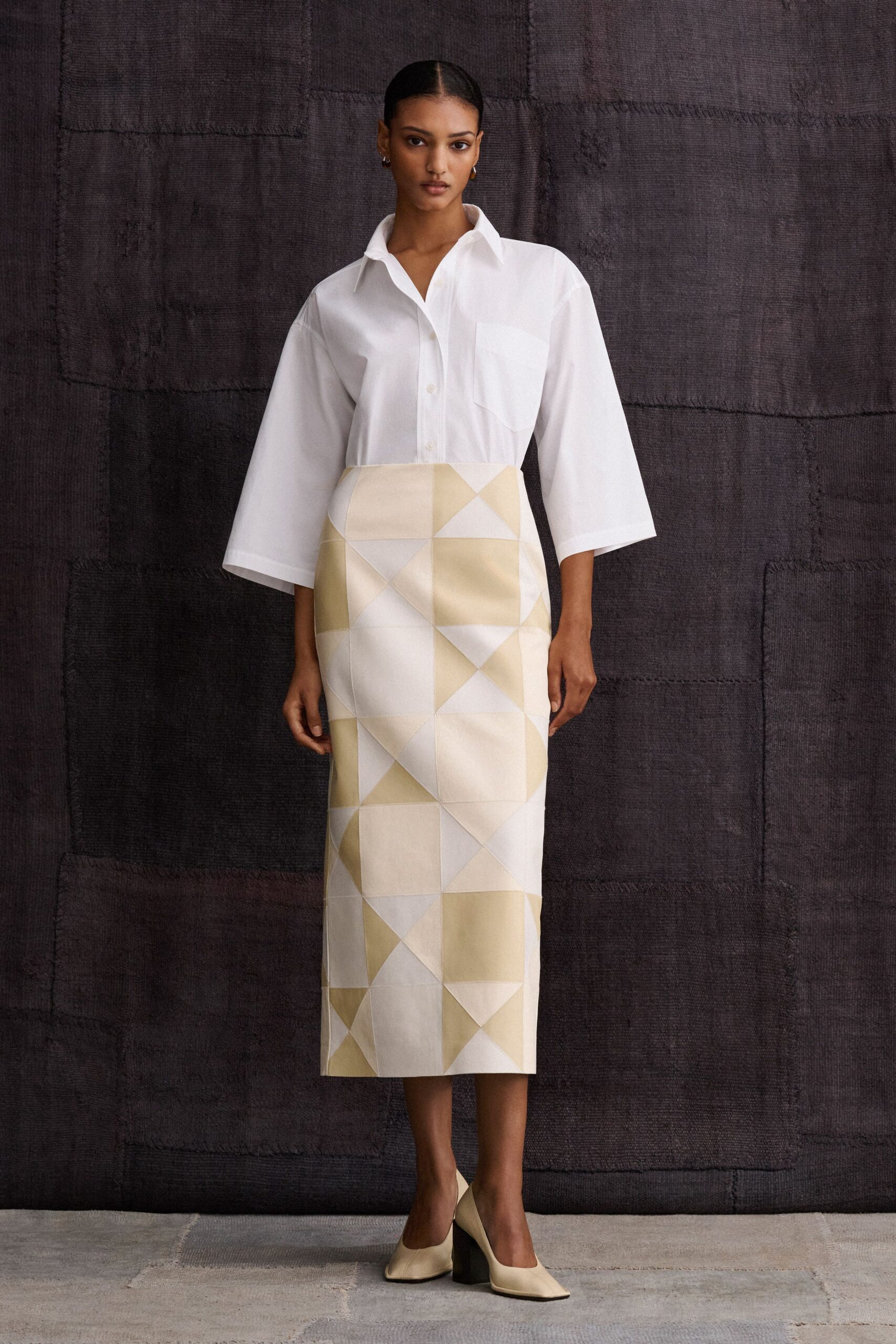 White cotton shirt with broad three-quarter sleeves and a skewed chest pocket. Cream midi pencil skirt pieced from geometric satin panels, seams crisply topstitched. Ivory pumps with squared toes and chunky black heels. For crossing a museum courtyard before the talk begins.