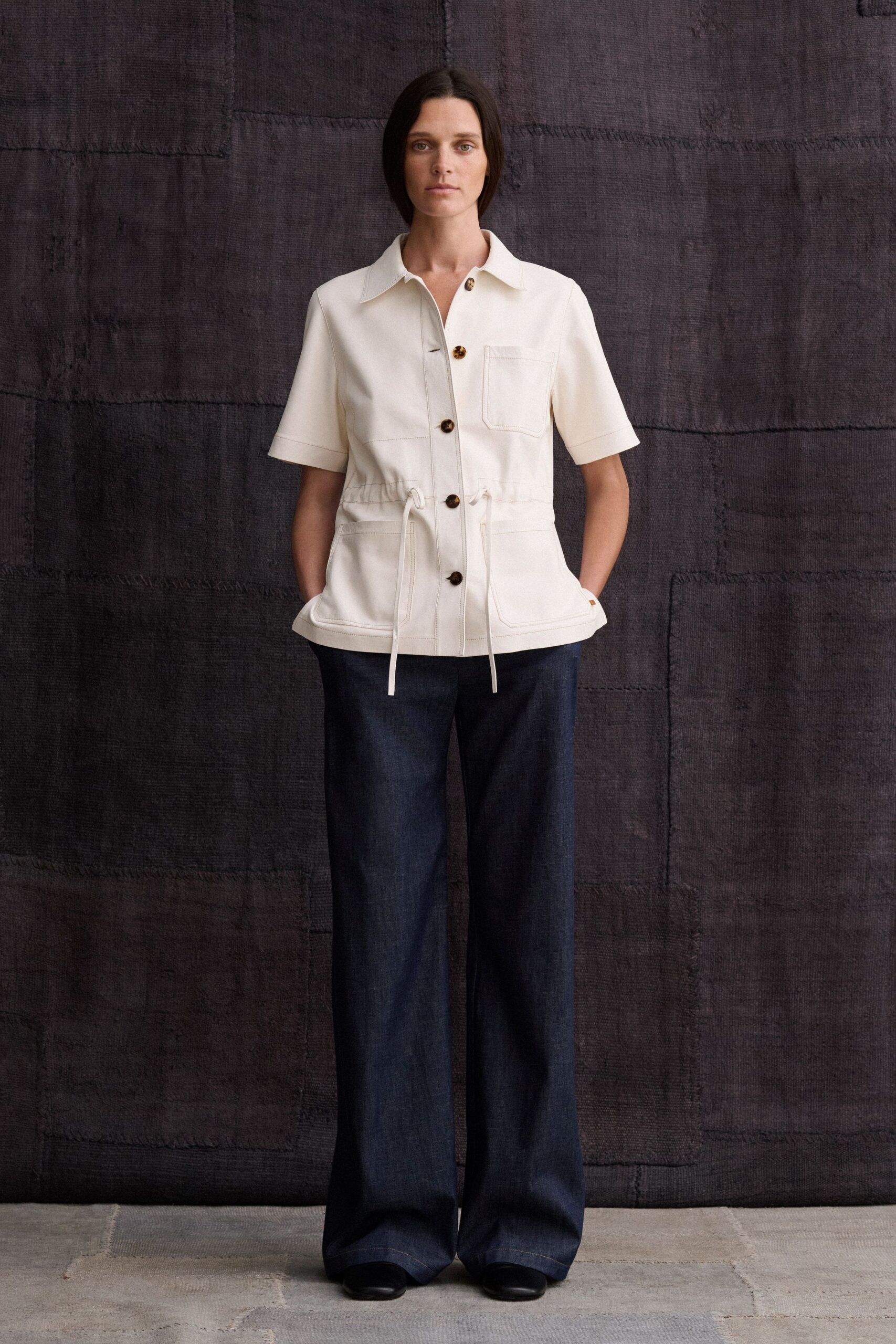 Cream twill shirt jacket with tortoiseshell buttons and a drawcord waist. Dark indigo wide-leg denim, pooling lightly over the shoes. Black leather round toes just peeking from the hem. For a museum morning that turns into lunch on the terrace.