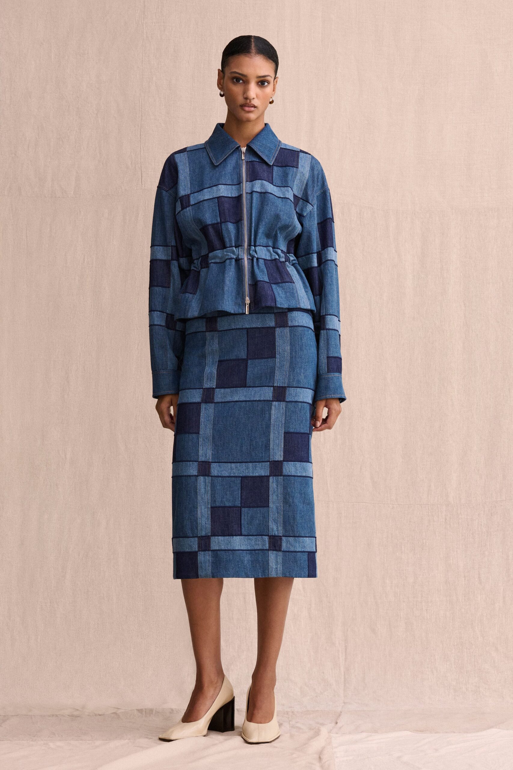 Patchwork denim jacket in indigo checks, zipped front cinched at the waist. Matching pencil skirt, broad grid panels cut from shifting blue washes. Cream leather pumps with blunt square toes and black block heels. Small gold hoop earrings. For a weekday meeting in a building with linen walls and no air-conditioning.