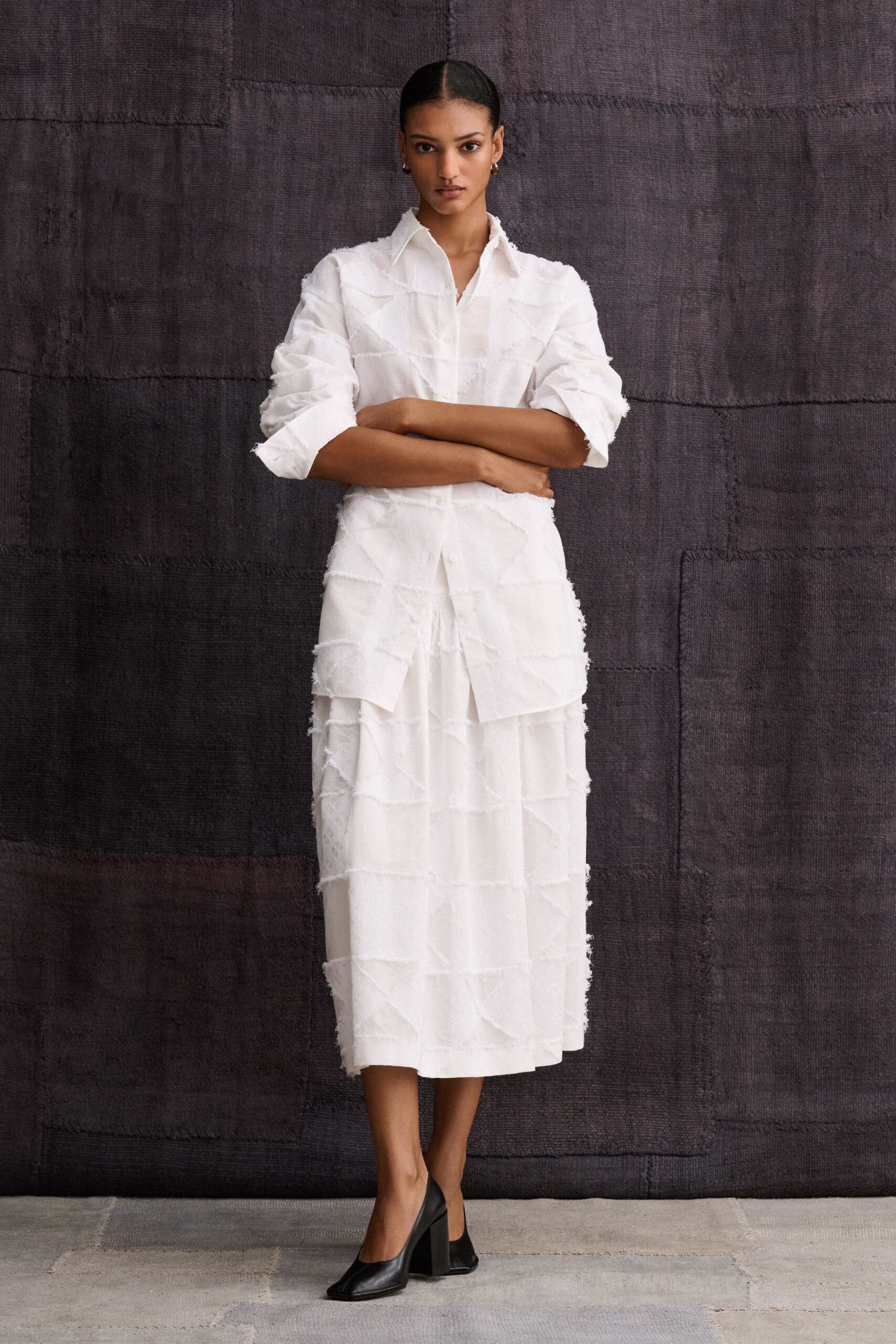 White shirt in gauzy open weave, raw horizontal bands lifting from the surface. Matching midi skirt, tiered with frayed strips and a soft sheer underlayer. Black leather pumps with sharply squared toes and thick block heels. For crossing a stone courtyard before the ceremony starts.