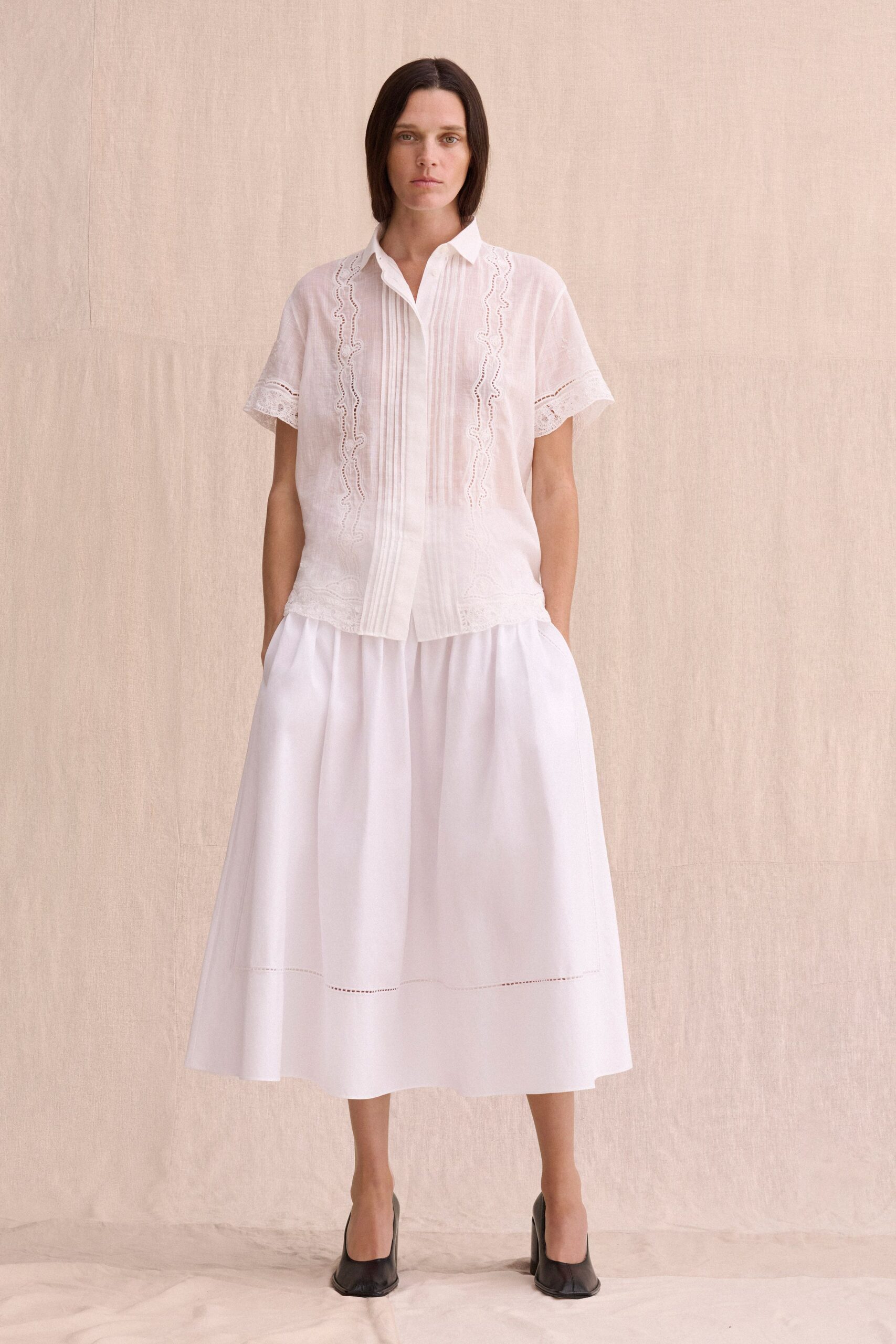 Sheer white cotton shirt with pintucks and scalloped broderie panels. Full white poplin midi skirt, dropped waist seam and ladder-stitch hem. Black leather pumps with squared vamps. Sunday rental house breakfast, curtains moving and nobody dressed for noon yet.