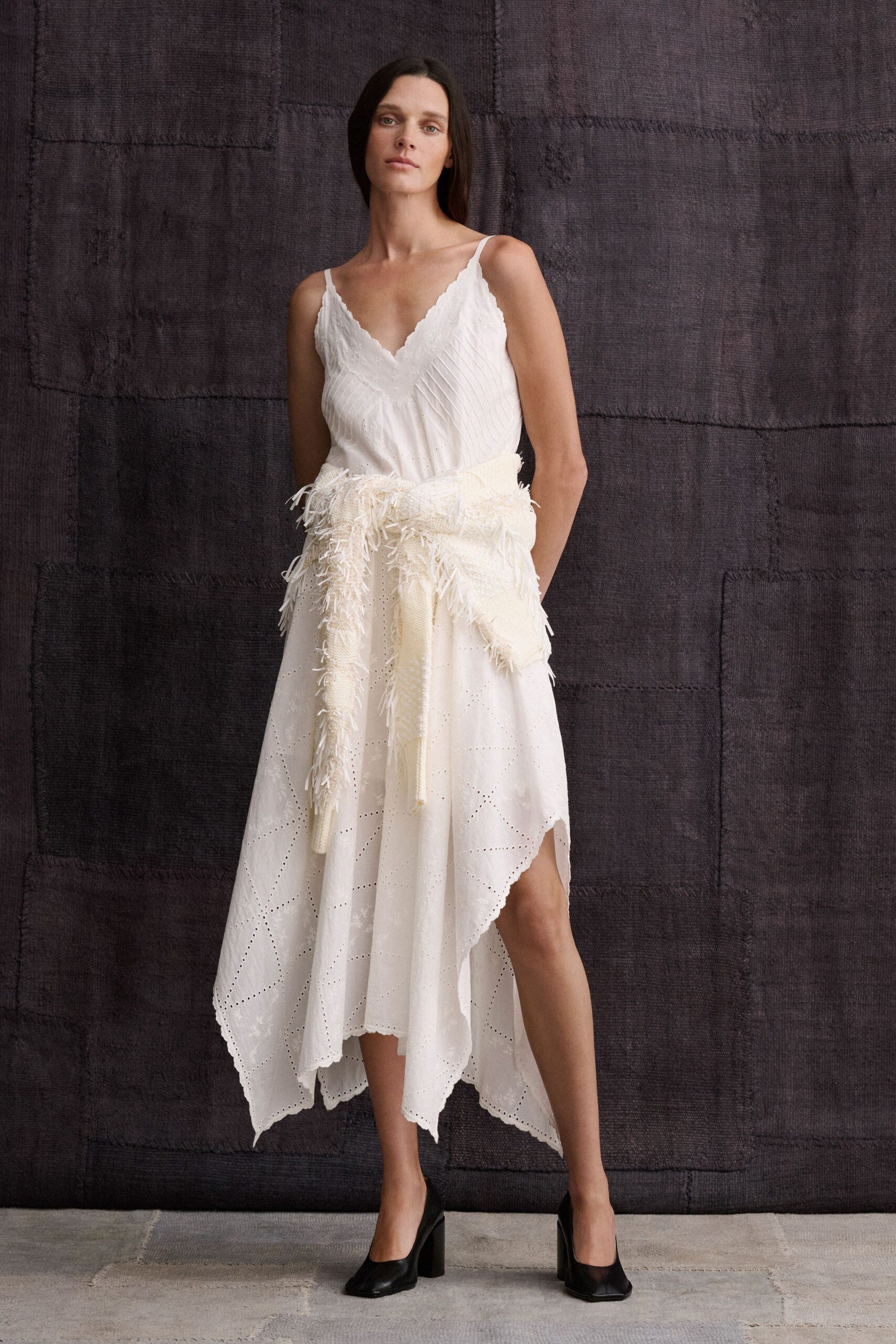Ivory slip dress with pintucked bodice, scalloped lace V and eyelet panels. Cream open-knit shawl knotted at the waist, shaggy fringe shedding outward. Black leather pumps with squared toes and thick block heels. Walking across a stone courtyard after the ceremony, bouquet already set down.