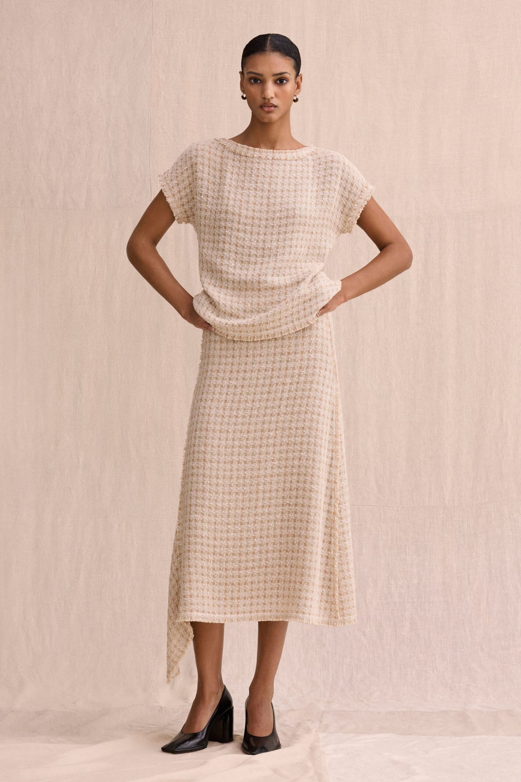 Cream open-weave bouclé top with frayed cap sleeves and a rolled neck. Matching midi skirt, softly flared with an uneven handkerchief point at one side. Black leather pumps with squared vamps and thick block heels. Small gold hoops. Gallery lunch in a converted house, white wine before the speeches start.