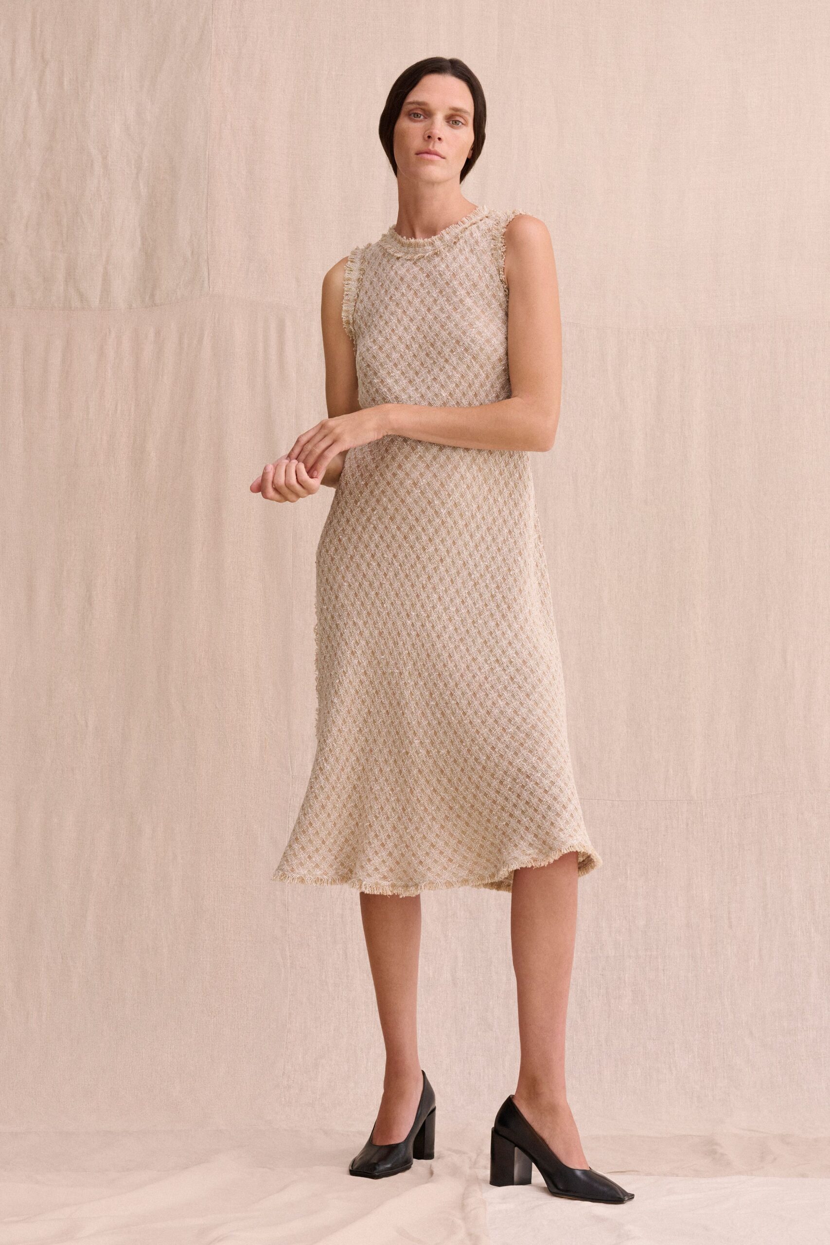 Oat boucle shift dress in a loose diamond lattice, fringed at neck and armholes. Black square-toe pumps with thick block heels. Museum atrium hush, just before the rain starts on the skylight.