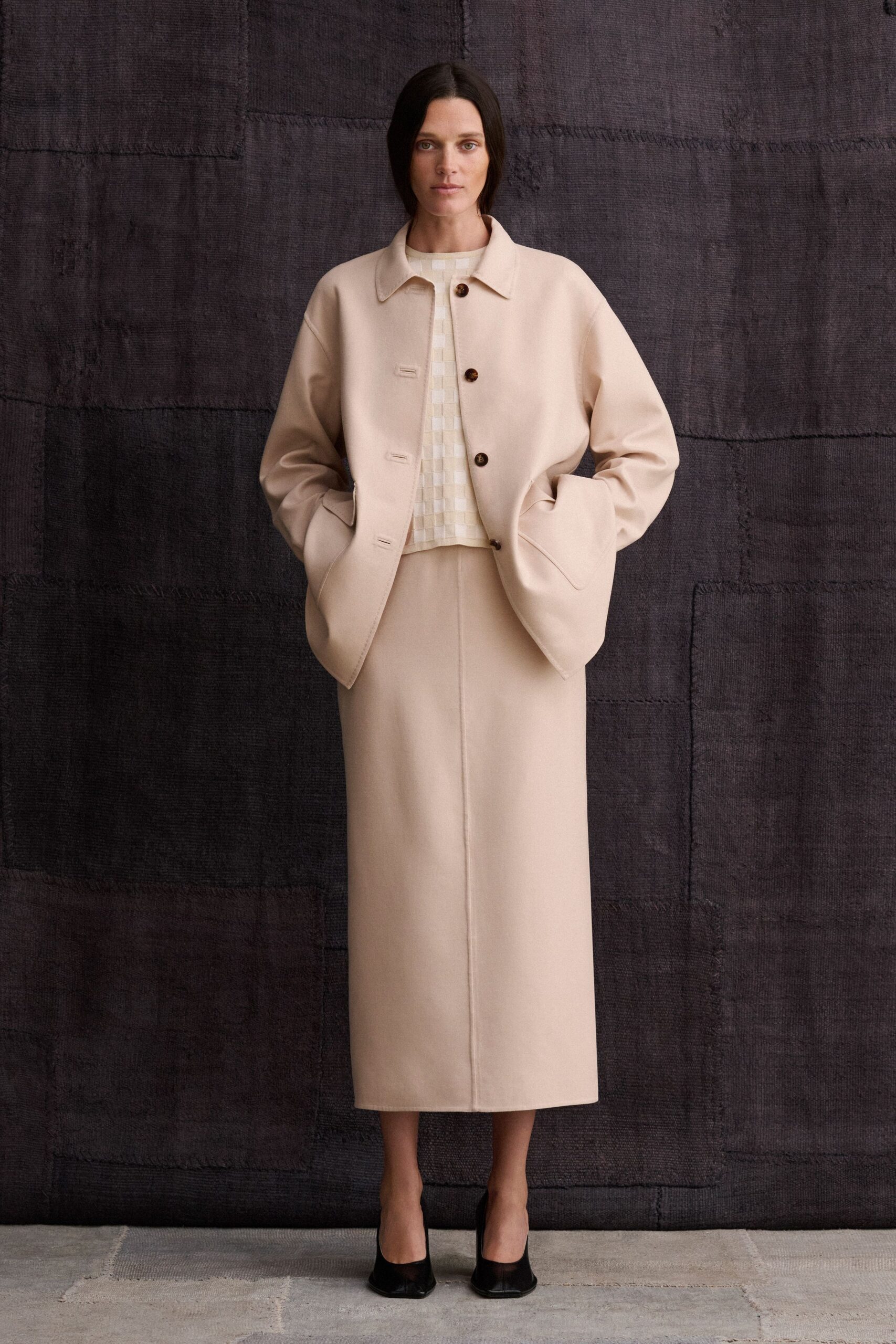 Cream felted jacket with horn buttons and low welted pockets. Ivory checkerboard-woven shell peeking from the open placket. Long column skirt in matching felt, center seam running straight down. Black leather pumps with a narrow squared toe. Museum foyer at opening hour, before the heat reaches the stone.