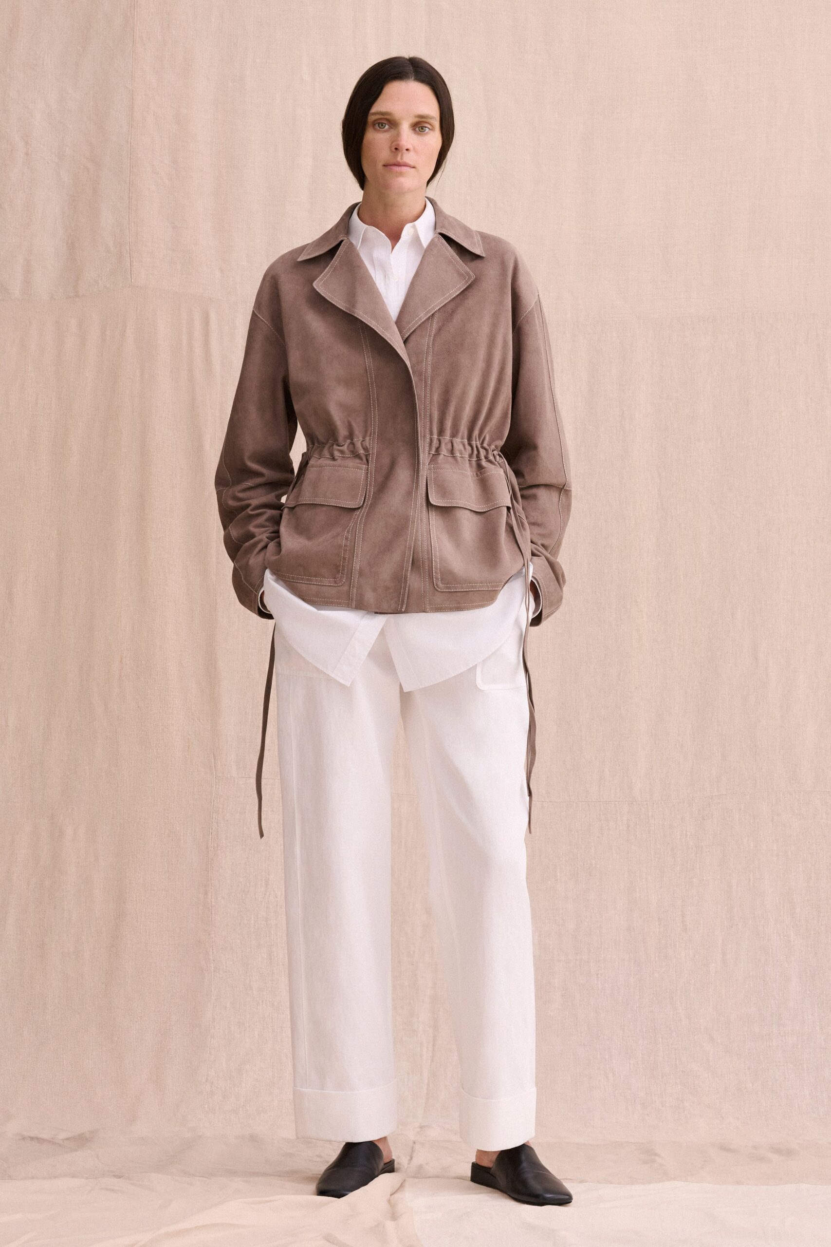 Taupe suede jacket with a ruched drawstring waist and topstitched patch pockets. White cotton shirt hanging long below the hem, collar kept crisp. Chalk wide-leg trousers with deep turned cuffs. Black leather mules with squared vamps. For the quiet hour before a house viewing, windows already open.