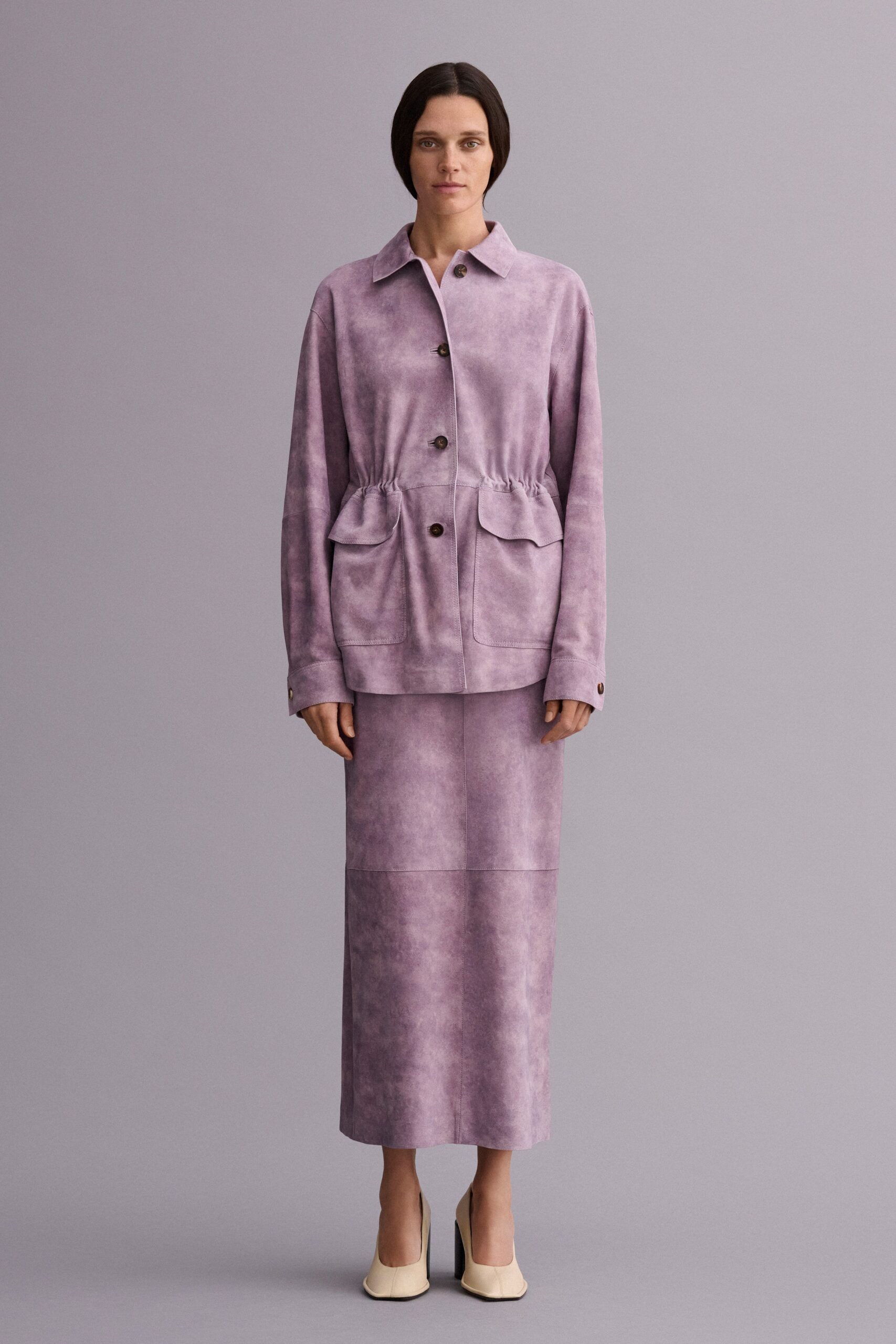 Lavender suede jacket washed cloudy pale and dark, waist softly ruched. Matching straight suede midi, paneled with a darker band near the hem. Cream pumps with squared toes and black flared heels. Museum atrium at opening hour, before the echoes fill with voices.