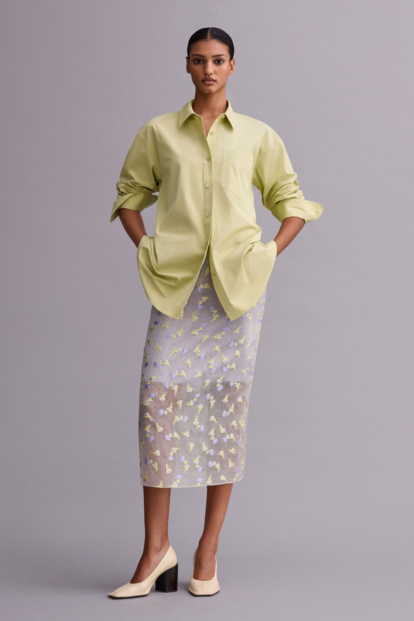 Pale pistachio cotton shirt, dropped shoulders and one curved chest pocket. Sheer crinkled midi skirt scattered with tiny chartreuse and periwinkle sprigs. Cream pumps with squared toes and glossy black block heels. For crossing a museum courtyard just after the rain stops.