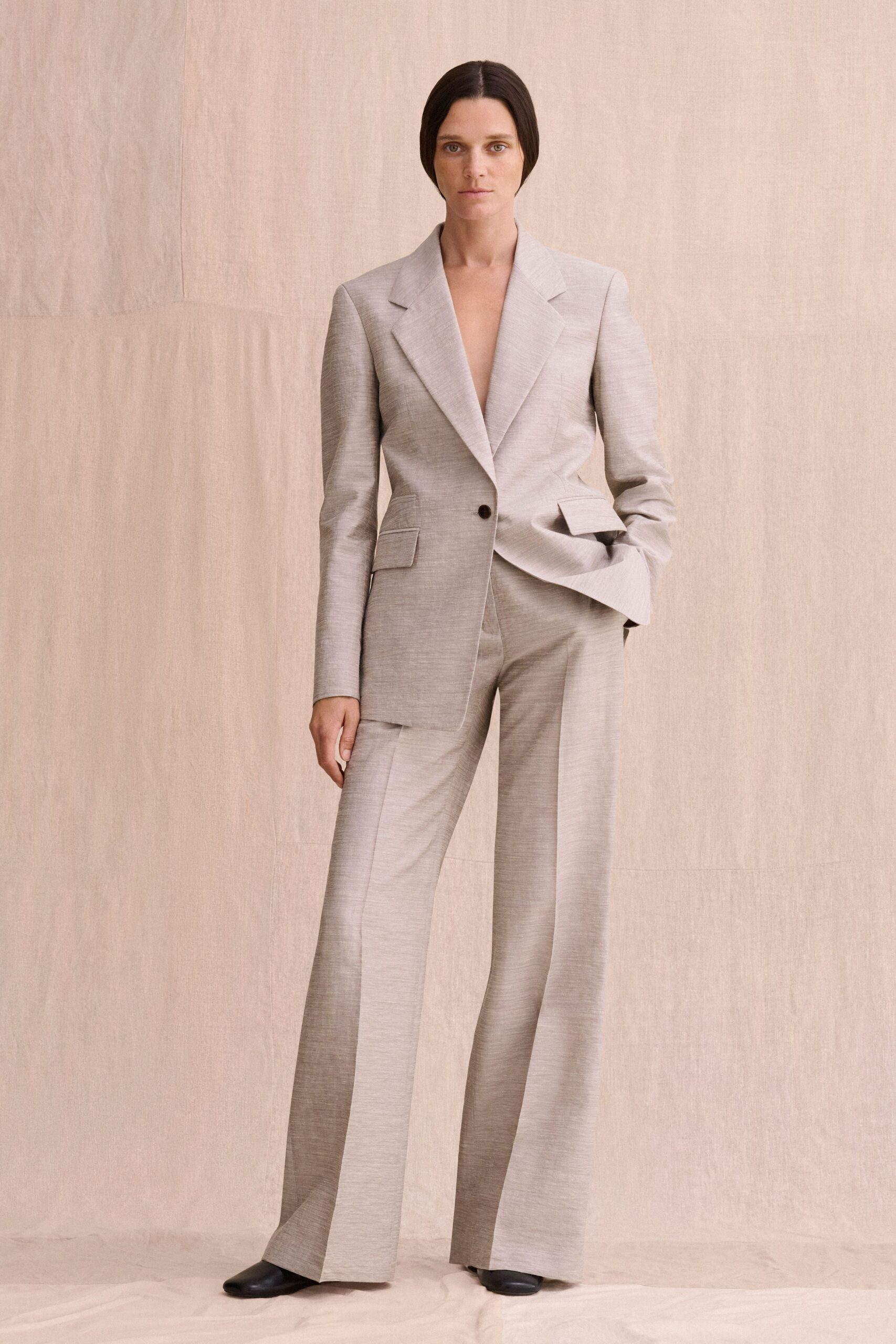 Pale greige suiting blazer with a plunging lapel and one dark button. Matching wide-leg trousers in dry crosshatch weave, pressed to a knife crease. Black leather square-toe shoes, barely showing under the hem. For the first meeting in an empty apartment before any furniture arrives.