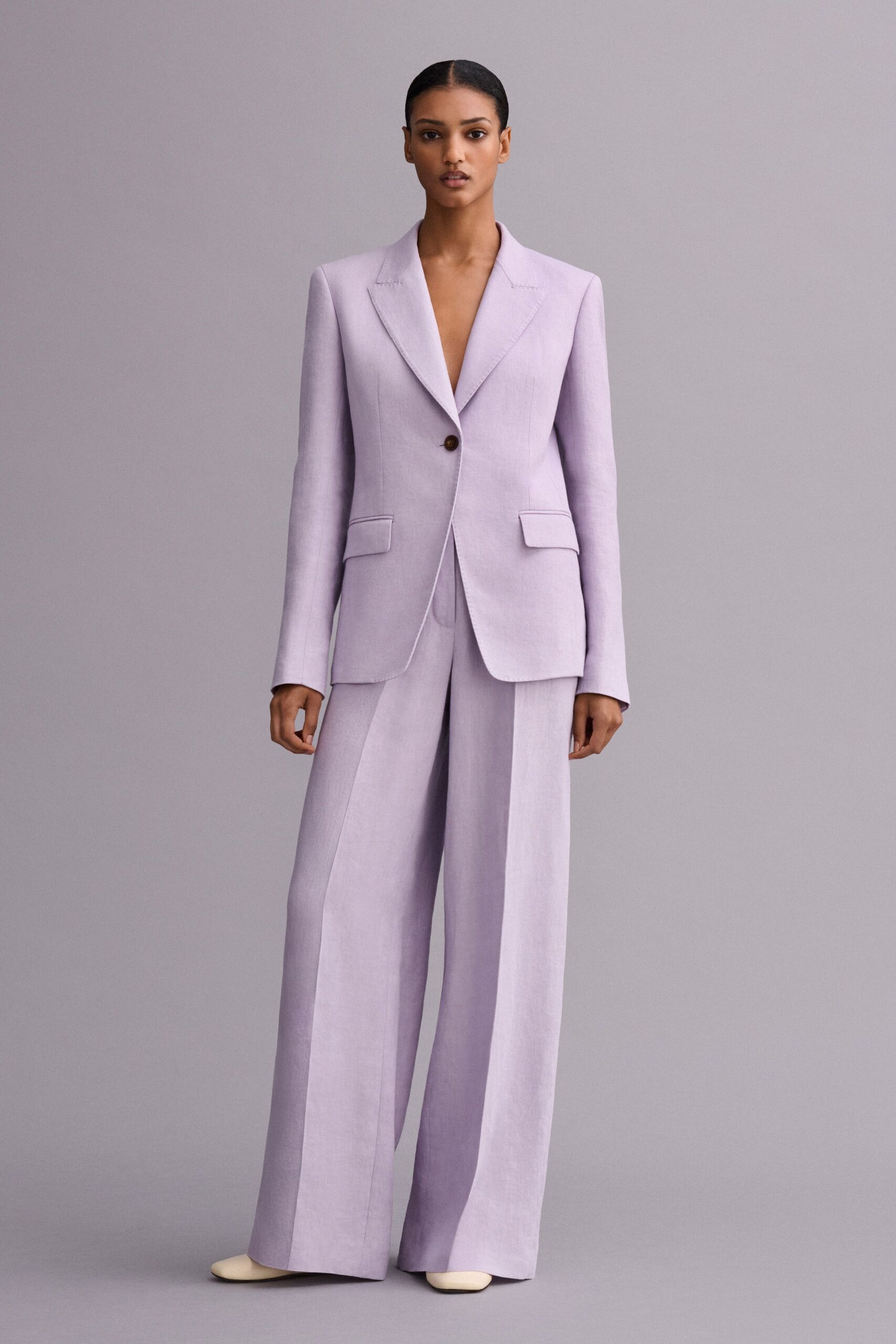 Pale lilac tailoring with strong shoulders and a single brown horn button. Matching floor-grazing trousers, pressed creases breaking over the shoes. Cream square-toe shoes peeking from the hem. For the courthouse wedding with only two witnesses and no flowers.