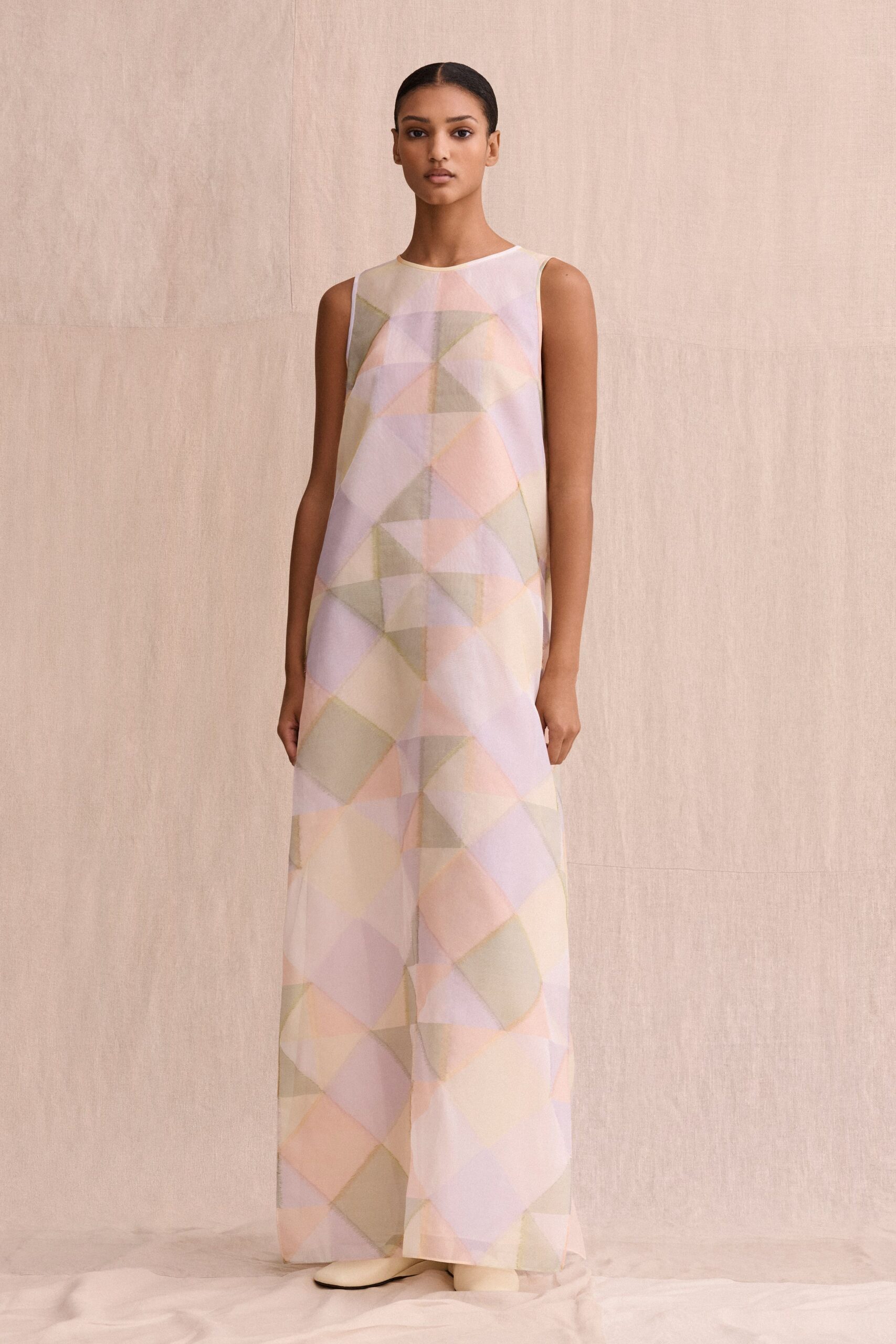 Floor-length sleeveless shift in sheer organza, washed with pastel geometric patchwork. Cream ankle boots with rounded toes peeking under the hem. Sunday morning in a rental house, curtains still turning the room milky.