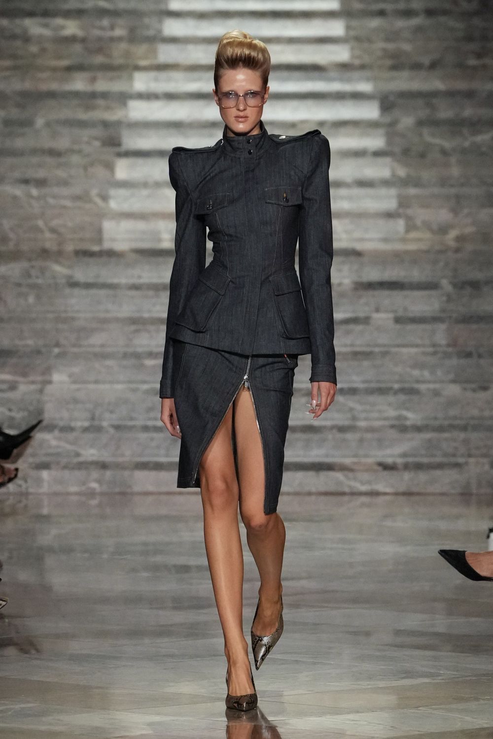 Oversized square aviator glasses with a pale smoke tint. Dark denim jacket cut razor-close, with pagoda shoulders and flap pockets. Matching denim skirt, front zip split riding sharply off center. Python pumps with pointed toes. For crossing a hotel lobby when the meeting has already gone hostile.