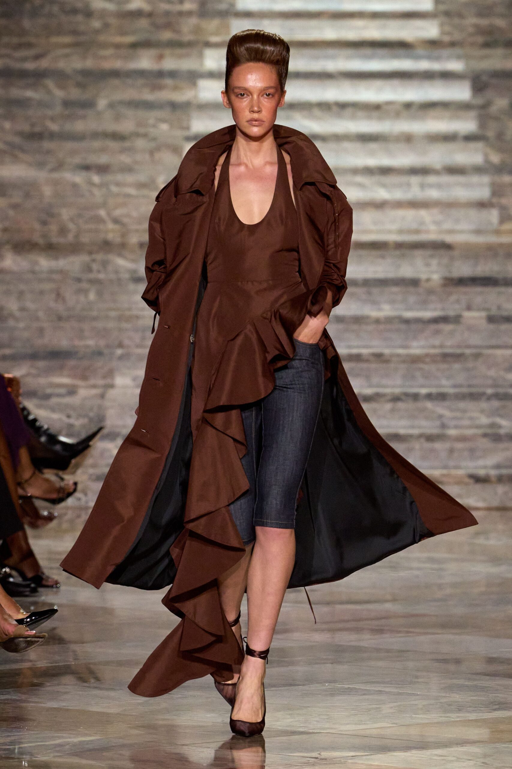 Chestnut floor-length coat with a blown funnel collar and black lining. Chocolate halter top cut low at the scoop neck, one side cascading ruffles. Dark indigo denim cycling shorts with pale topstitching. Brown mesh pumps with slim wraparound ankle ties. For crossing a museum atrium when the storm has just broken outside.