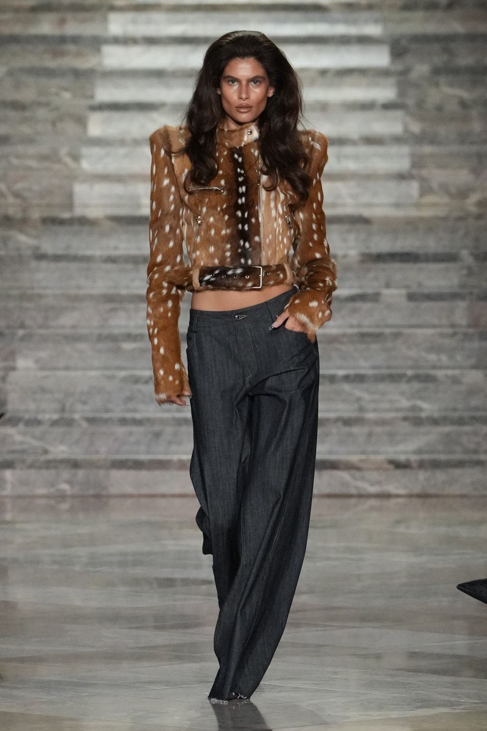 Cropped fawn-hide jacket with white spots and jutting shoulders. Matching hair-on hide belt with a square metal buckle. Charcoal wide-leg trousers in dry suiting, hems dragging into threads. Whatever you wear to cash a settlement check in a mountain town.