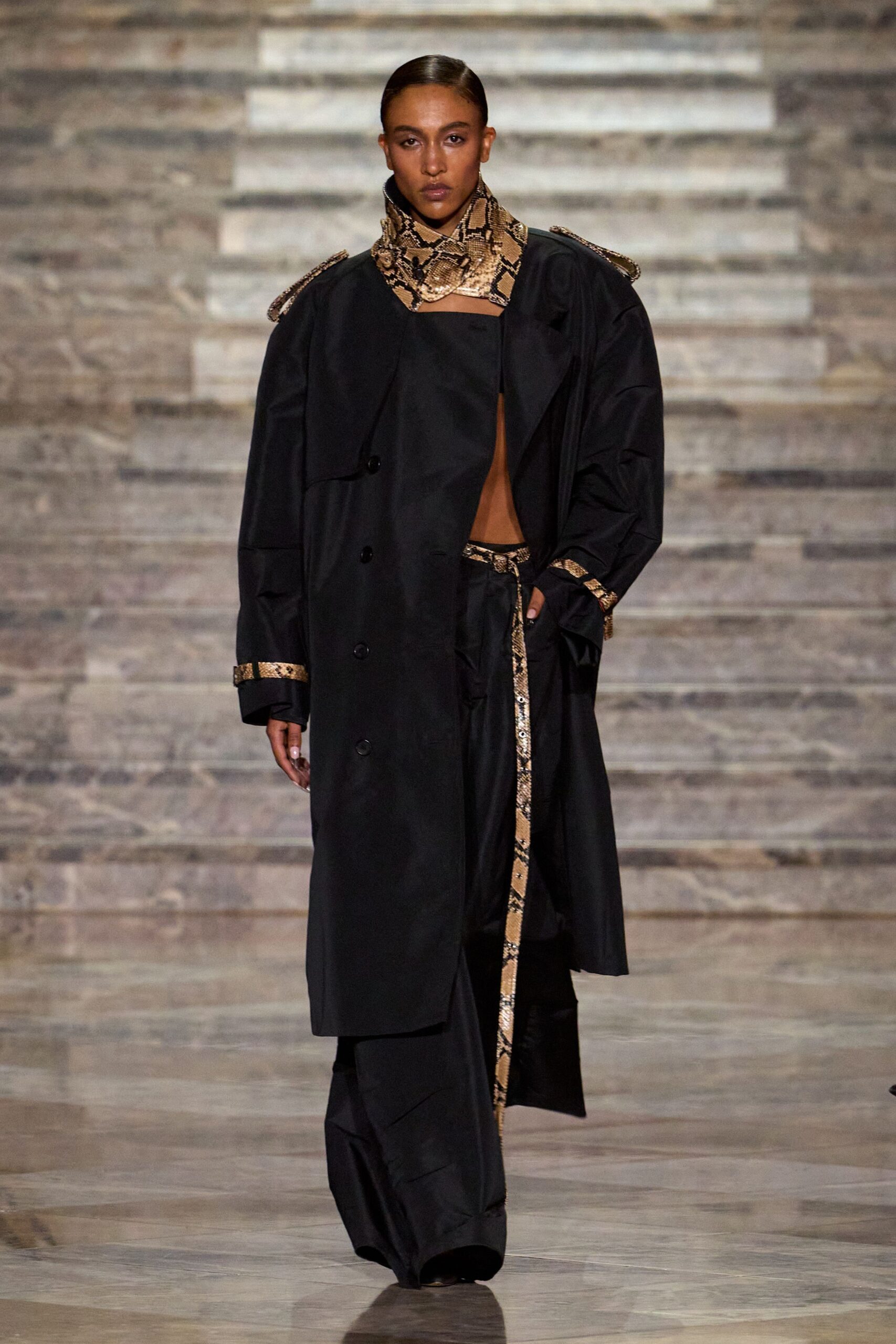Tan python scarf buckled high at the throat, one pointed tab overlapping. Black trench cut oversized, python tabs circling cuffs and epaulettes. Wide black trousers with a dangling python belt strip down the front. Walking into a hotel atrium still damp from rain, keycard already in hand.