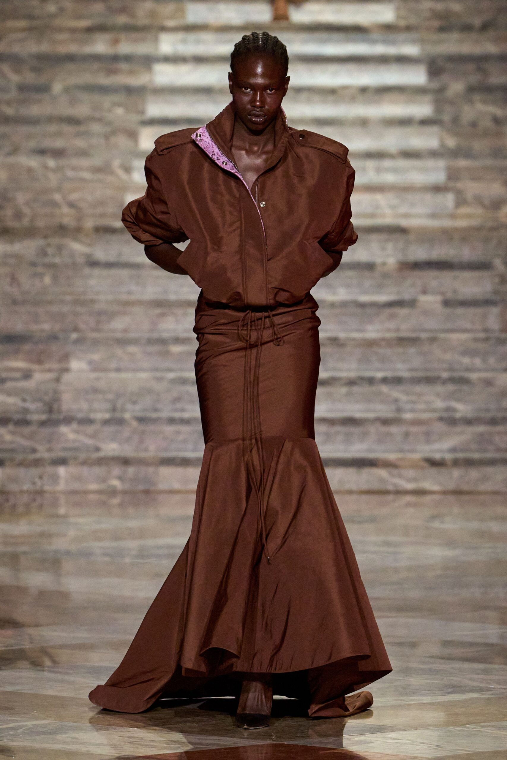 Chocolate zip-front bomber in crisp twill, pink sequins glinting inside the collar. Matching fishtail skirt, drawcords dangling from a dropped waist seam. Square-toe boots swallowed by the pooling hem. For crossing a stone courtyard just after the rain stopped.