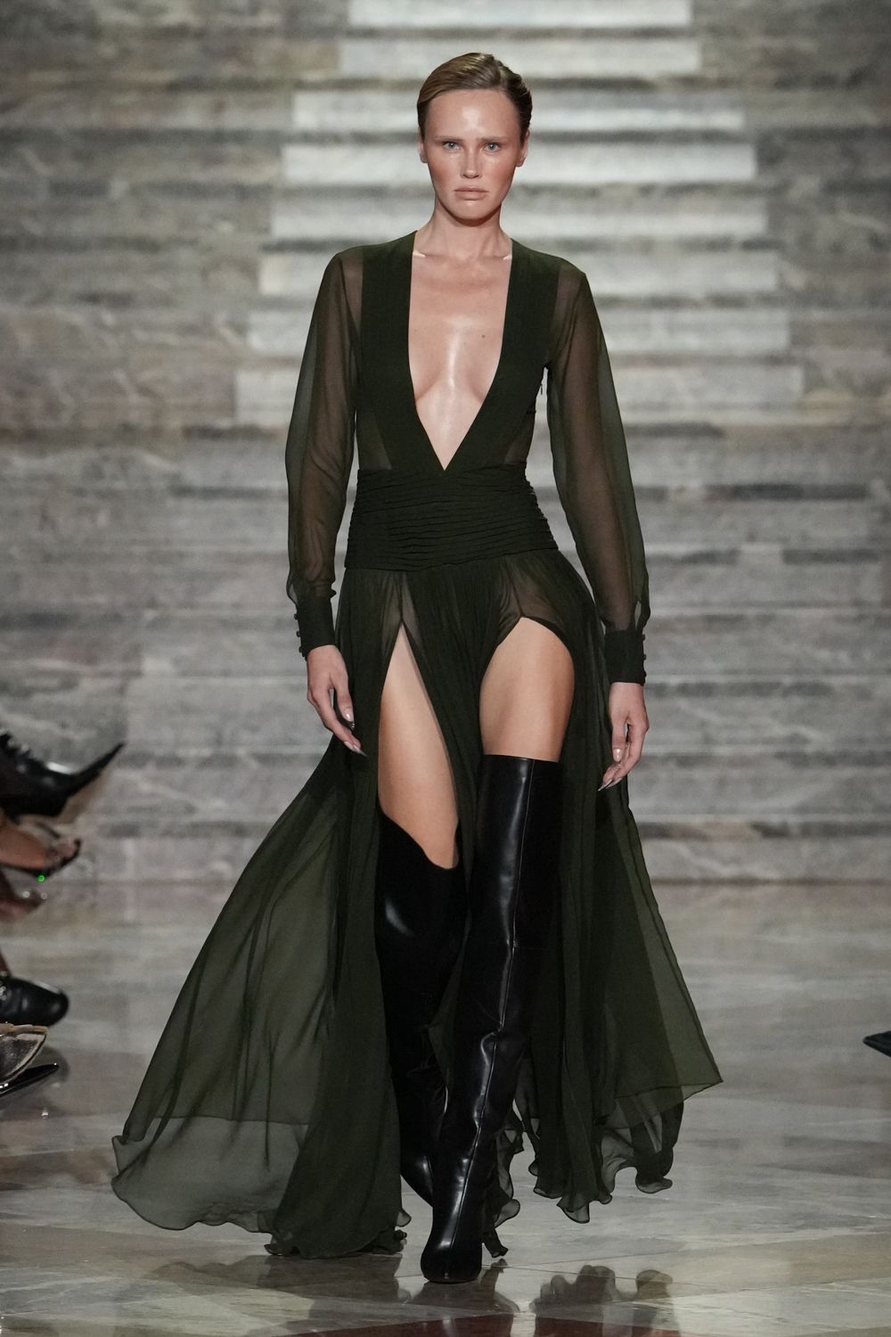 Deep olive chiffon gown with a plunging V and ruched waist panel. Black thigh-high leather boots, hard shine against the floating split skirt. Whatever you wear home when the hotel bar turned into dawn.