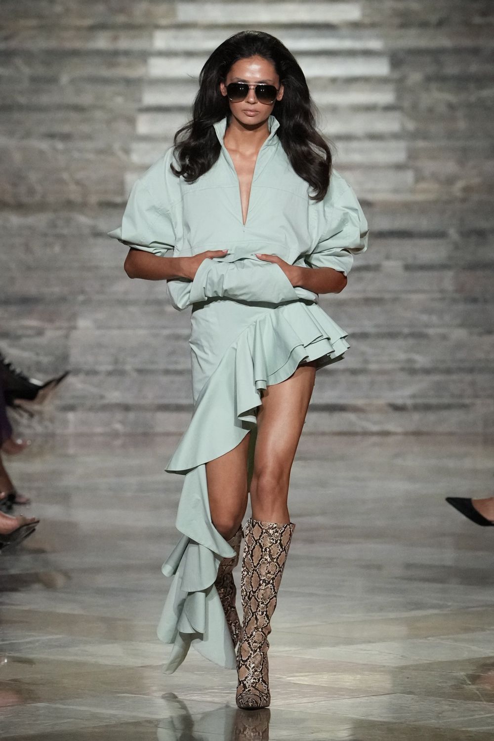 Oversized smoke aviators with a thin metal bridge. Powder-mint poplin dress, half-zip plunged open and one ruffle tail sweeping low. Matching opera gloves in the same crisp cloth. Python knee boots, softly slouched through the shaft. Walking into a hotel lobby still carrying the sea breeze.