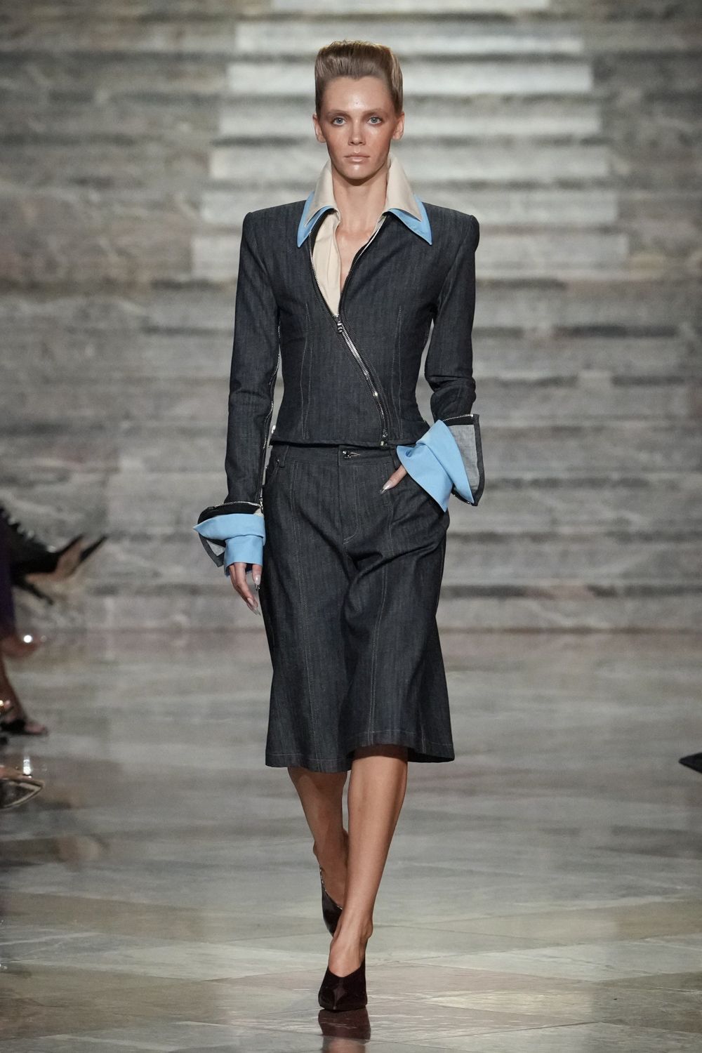 Dark denim jacket cut close, closing on a slanted silver zip. Cream shirt with oversized powder-blue collar and turned-back cuffs. Dark denim culottes with pale topstitching and a low curved fly. Sharp black pumps. For the Monday presentation where the air-conditioning is always too cold.