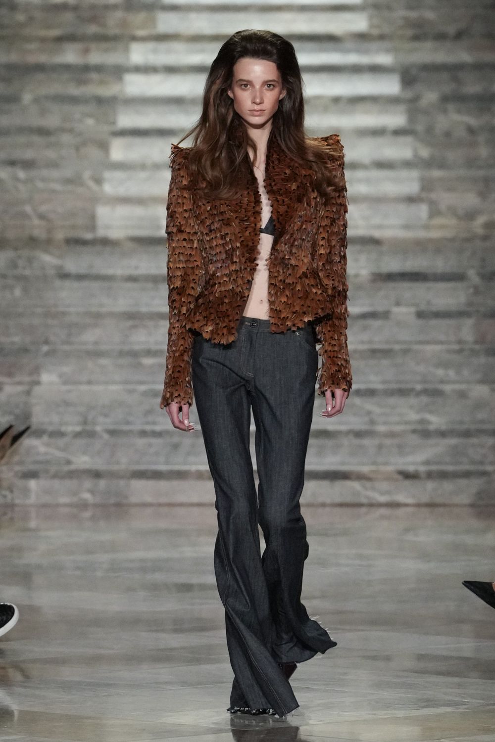 Chestnut jacket covered in dense cut leather fronds, hem left jagged. Tiny black triangle bra peeking through the open front. Charcoal bootcut trousers with a low rise and pooling break. Dark pointed toes barely visible under the dragging hem. For crossing a hotel lobby after midnight with your room key in hand.