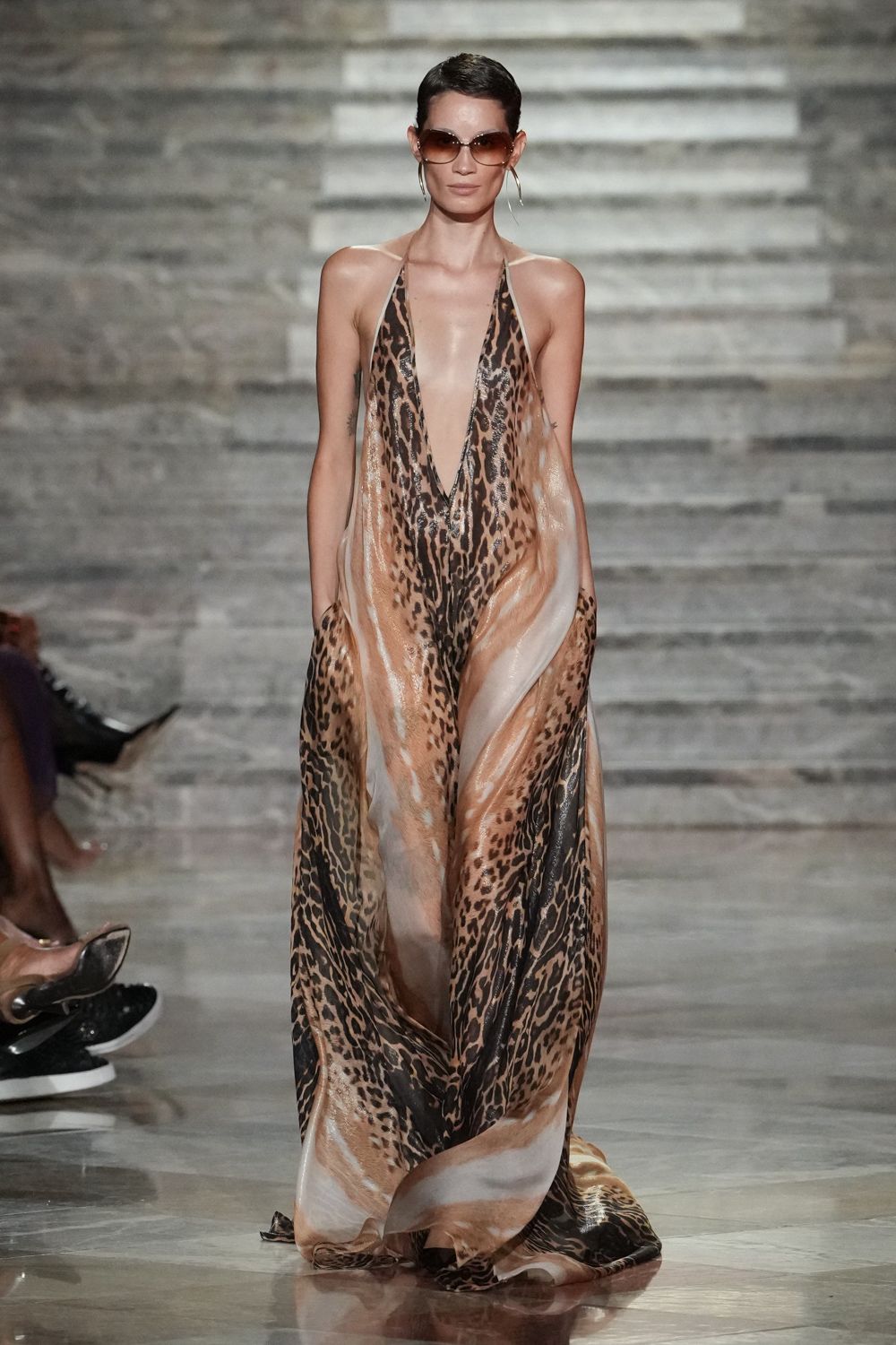 Oversized brown gradient lenses with a softly squared rim. Thin silver hoops, one swinging lower than the jaw. Plunging halter gown in liquid leopard satin, side drapes opening with each step. Hotel pool after midnight, the tiles still warm underfoot.