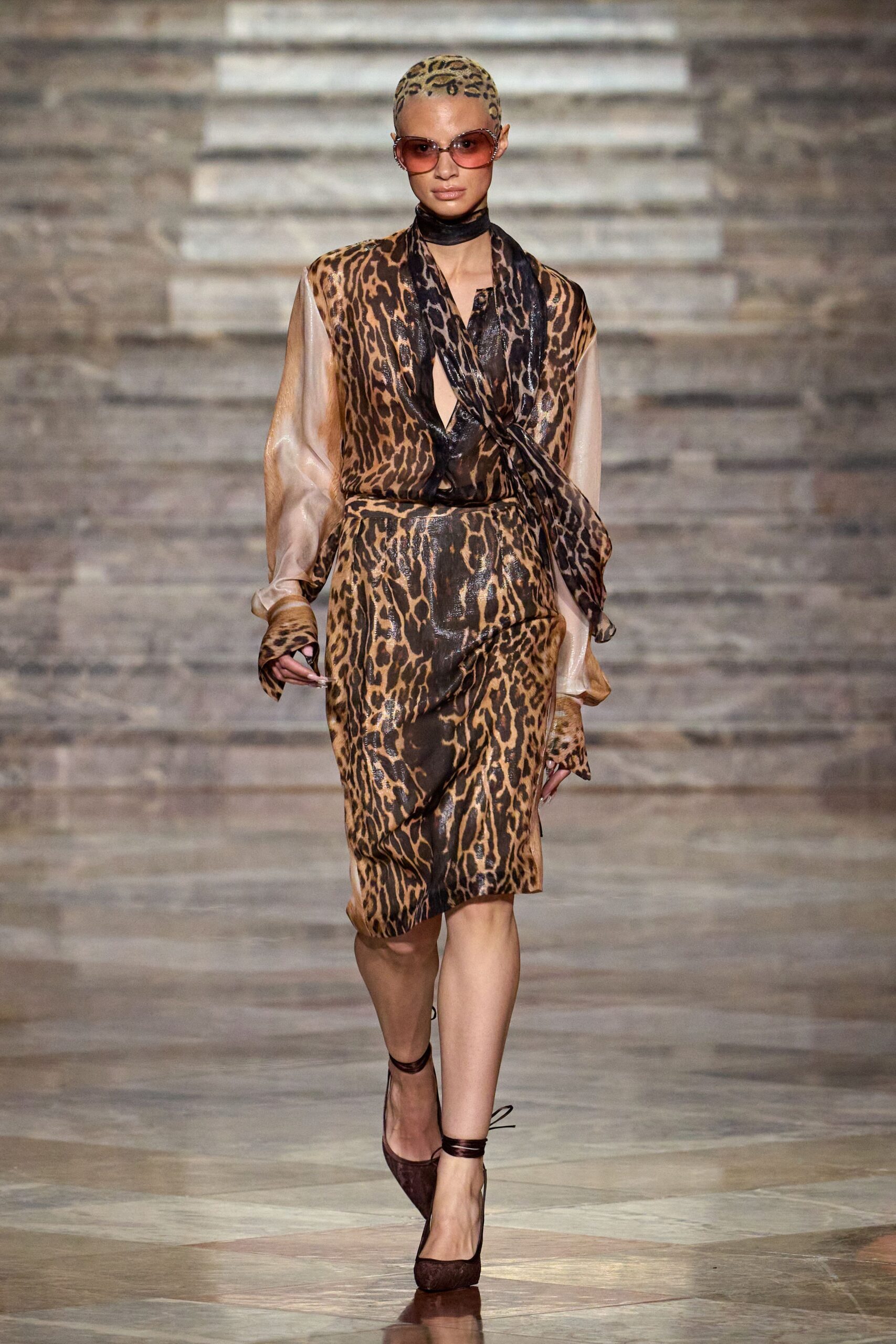 Rose-tinted oversized frames with a soft square fade. Leopard scarf wrapped tight over the cropped head. Leopard satin dress with sheer champagne sleeves and a scarf-tie plunge. Burgundy pumps laced at the ankle. Hotel terrace at noon, ordering something cold before the table arrives.