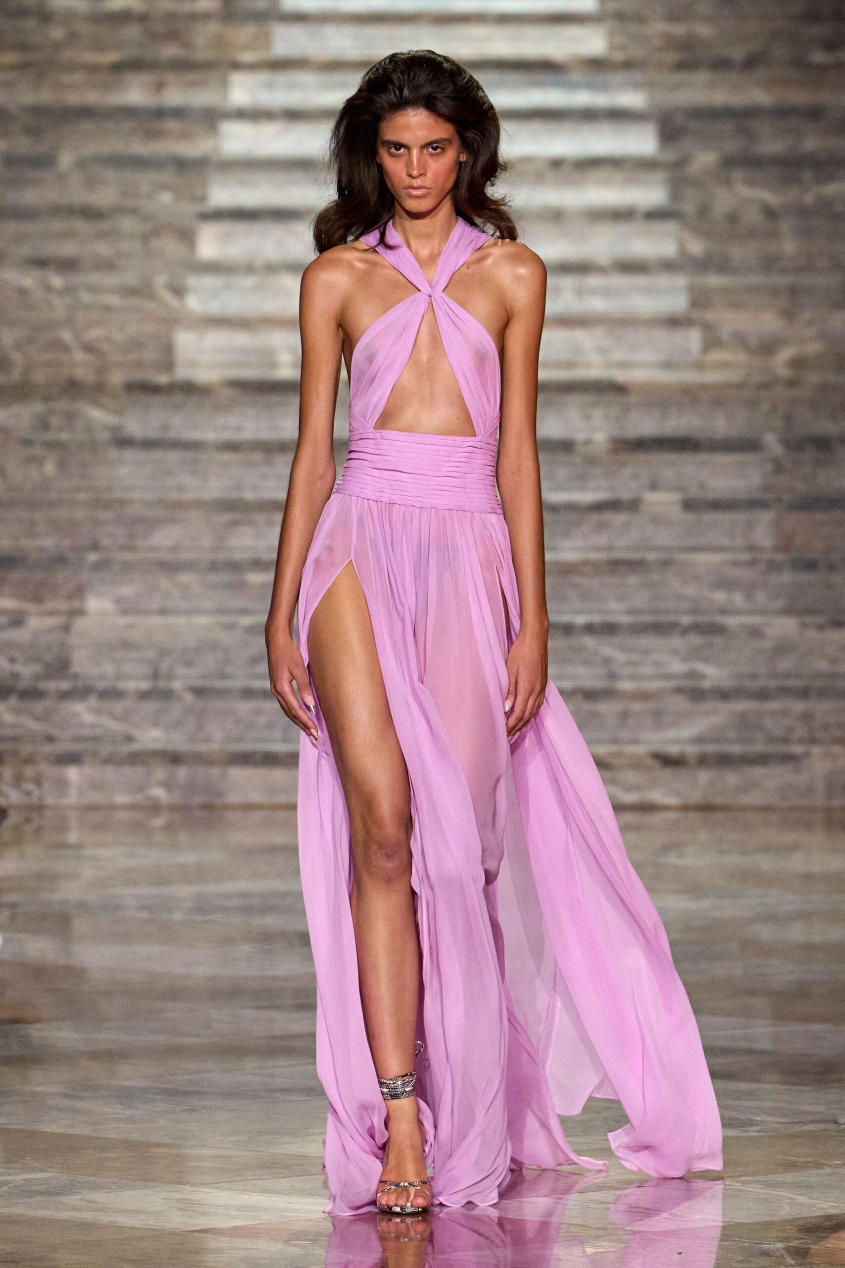 Lilac chiffon halter gown twisted at the neck, ruched waistband, thigh-high slit. Silver barely-there sandals with a coiled anklet strap catching the light. For crossing a hotel terrace just after the pool party emptied out.