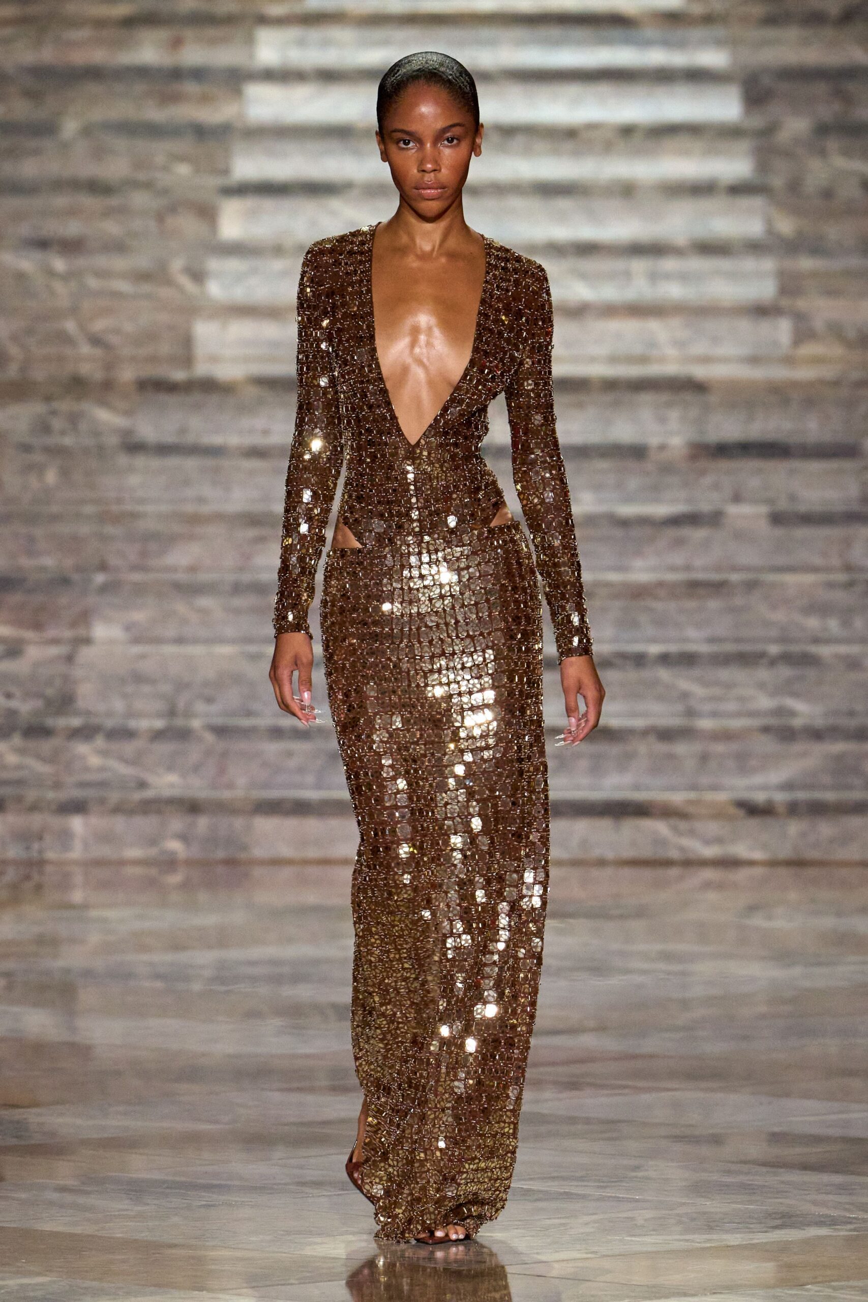 Bronze mirror-tile column gown with a plunging V and hip cutouts. Barely there bronze sandals peeking from the pooled hem. Hotel ballroom after midnight, the staff are stacking chairs around you.