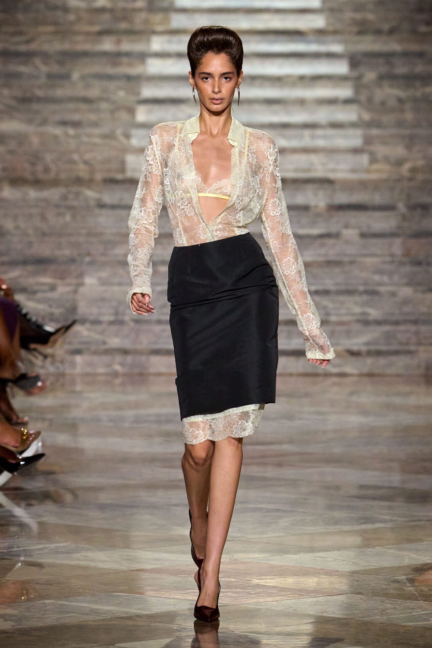Long silver hoops grazing the jaw. Pale lemon lace triangle bra visible through the plunge. Sheer ivory floral lace blouse, collar soft and slightly buckled. Black pencil skirt draped low at one hip, lace hem exposed. Oxblood pointed pumps in glossy patent. Walking into a hotel bar before dinner service has properly started.